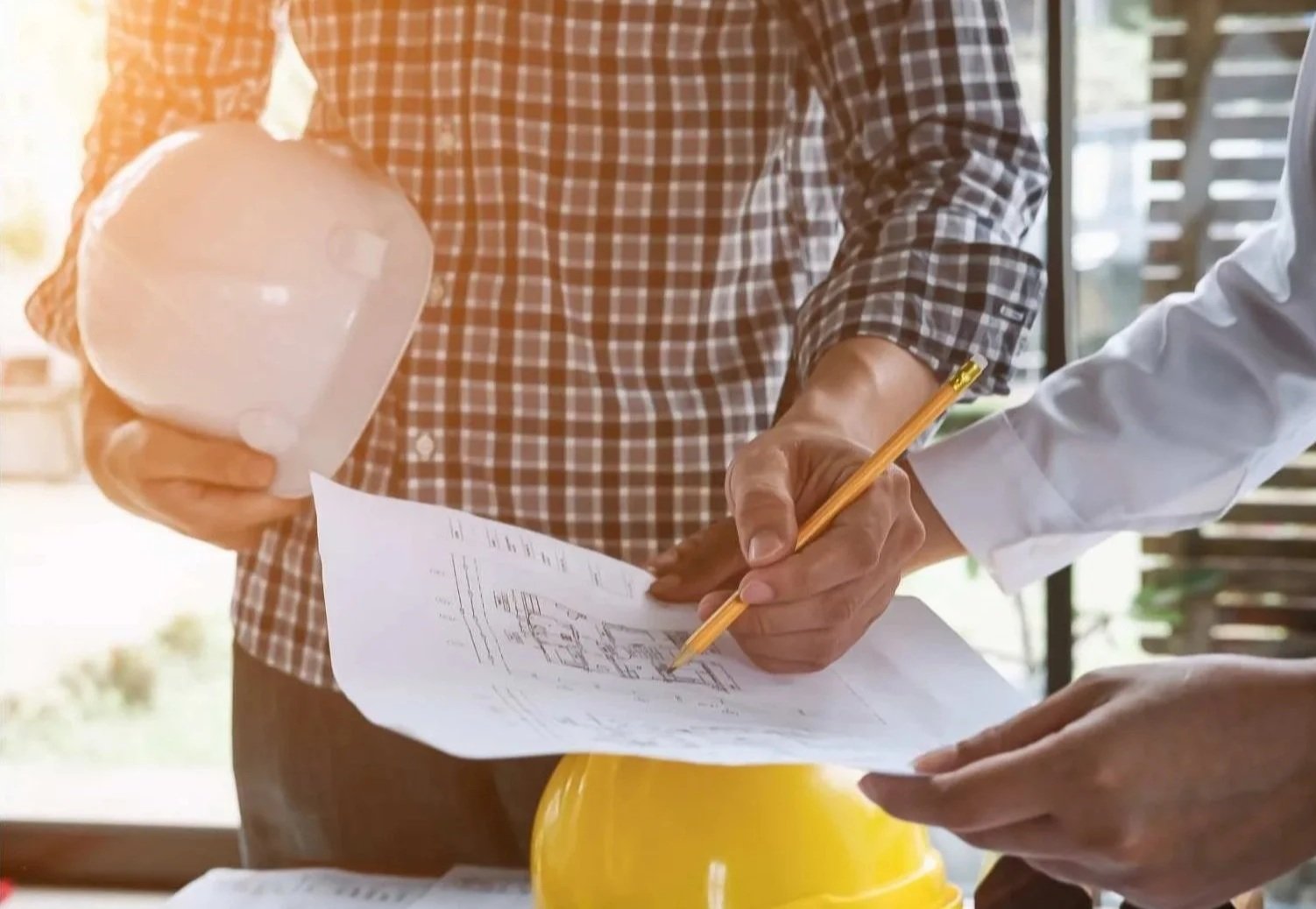 Two individuals review construction plans; one holding a safety helmet, the other writing on the plans.