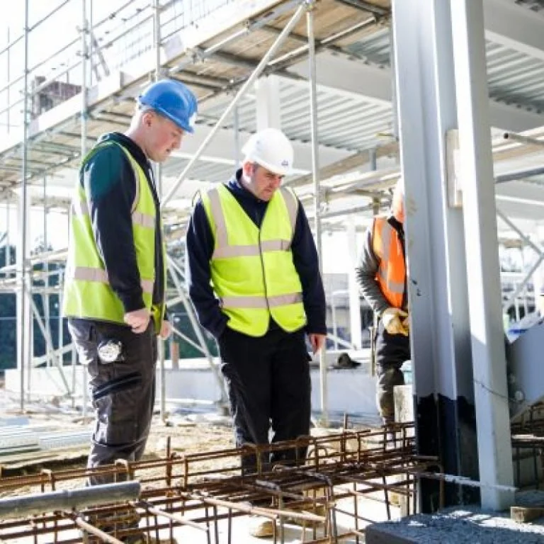 Three construction workers wearing safety vests and hard hats inspecting a construction site with steel reinforcement bars and scaffolding.