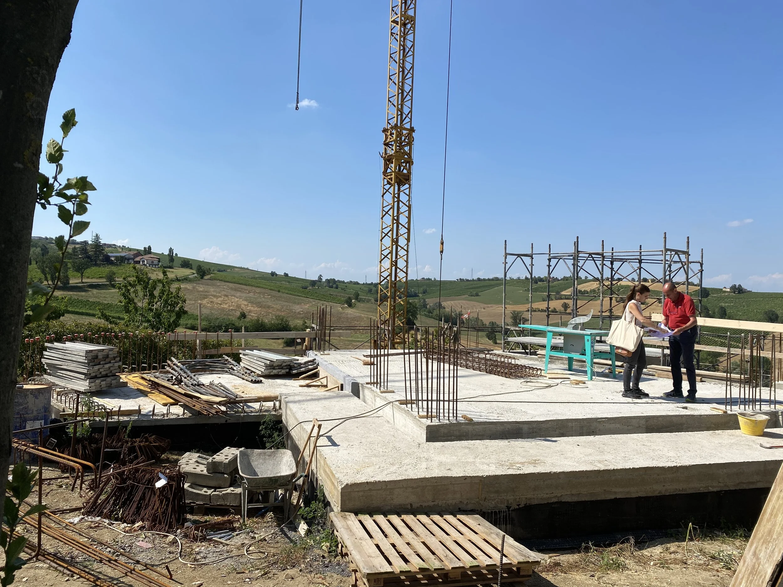 Construction site with concrete foundation, steel reinforcement bars, a yellow tower crane, and two people reviewing plans on a construction table in a rural area with green hills in the background.