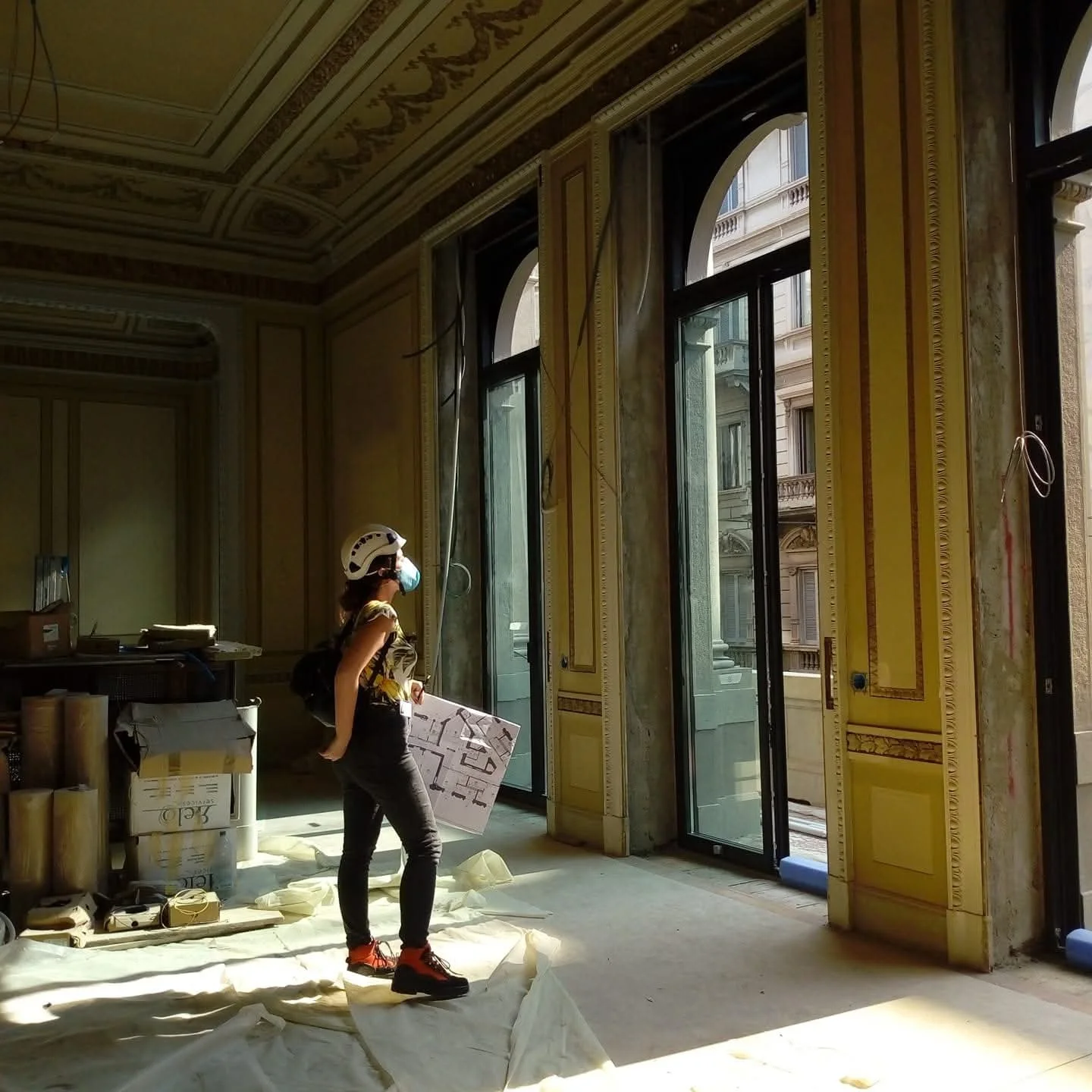 Architect wearing a helmet, face mask, and backpack inside a historic building under renovation, holding a floor plan, with large windows and ornate yellow walls.