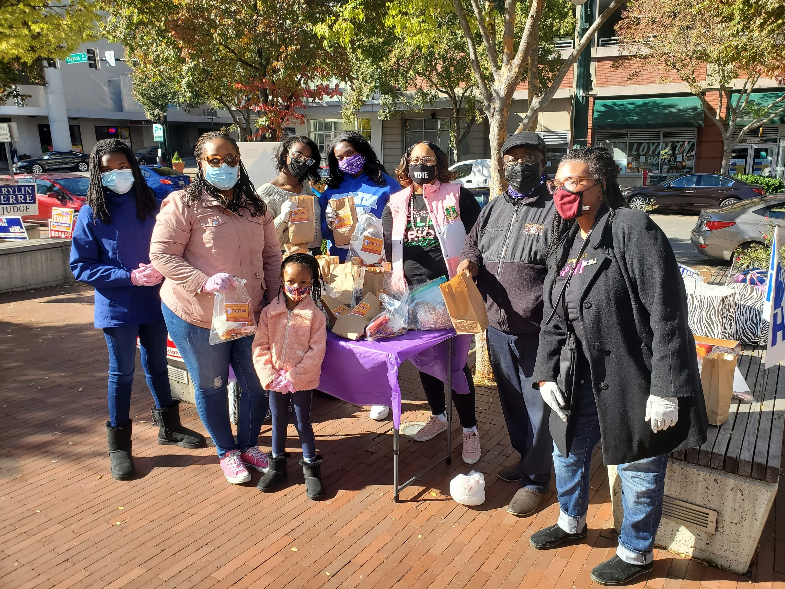 Group of seven people, mostly women, standing outdoors on a city street, wearing face masks, some holding paper bags, and near a table with more bags, with trees and storefronts in the background.