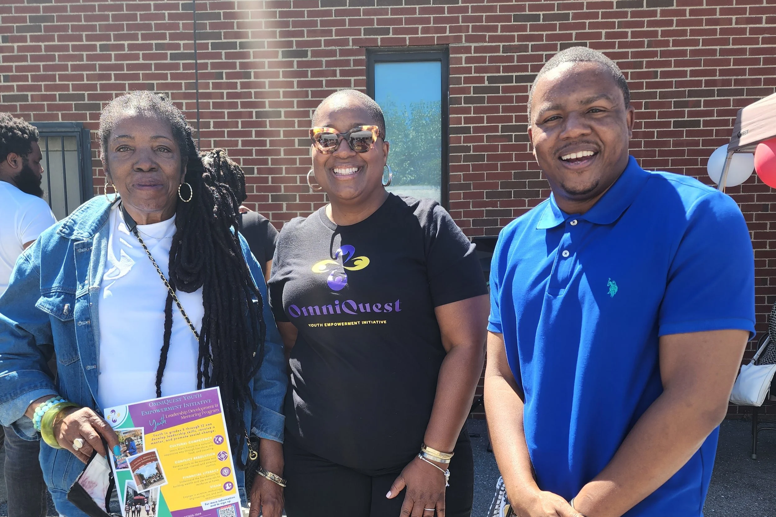 Three people standing together outdoors in front of a brick building, smiling for the photo. The person on the left is an older woman with long dark hair, wearing large hoop earrings, a white shirt, and a denim jacket. The person in the middle is a w
