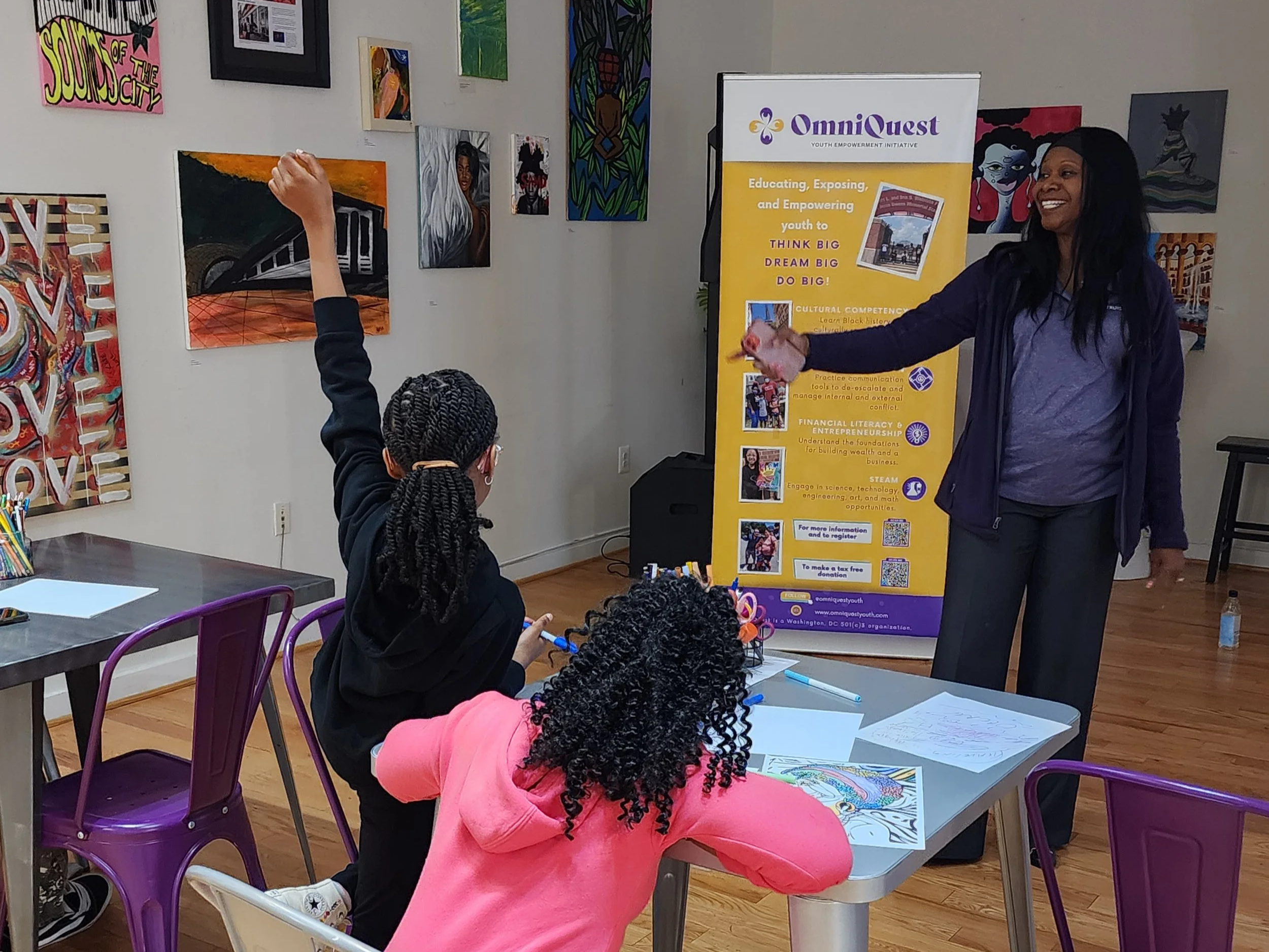 A classroom setting with children seated at tables, drawing or coloring on paper. A woman stands at the front near a yellow banner that reads 'OmniQuest' and 'Educating, Exposing, and Empowering youth to THINK BIG, DREAM BIG, DO BIG!' The walls are d
