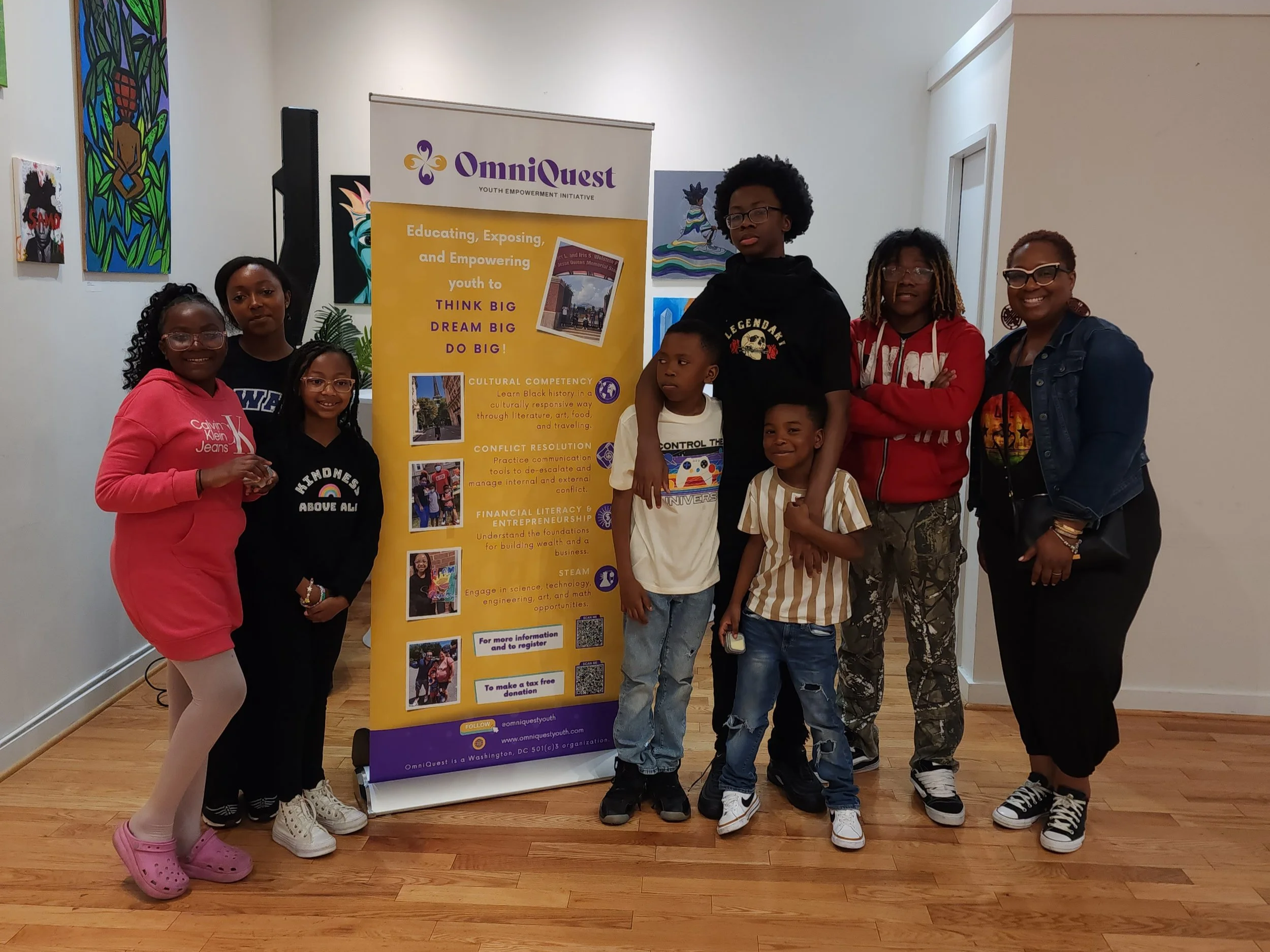 A group of seven young people standing in front of a colorful display and a yellow banner for OmniQuest Youth Empowerment Initiative, smiling and posing for a photo inside a gallery or community space.
