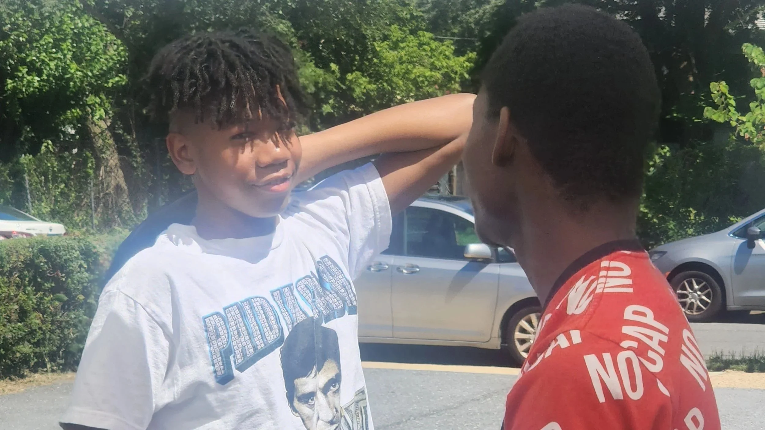 Two young boys having a conversation outdoors in a parking lot with cars and trees in the background.