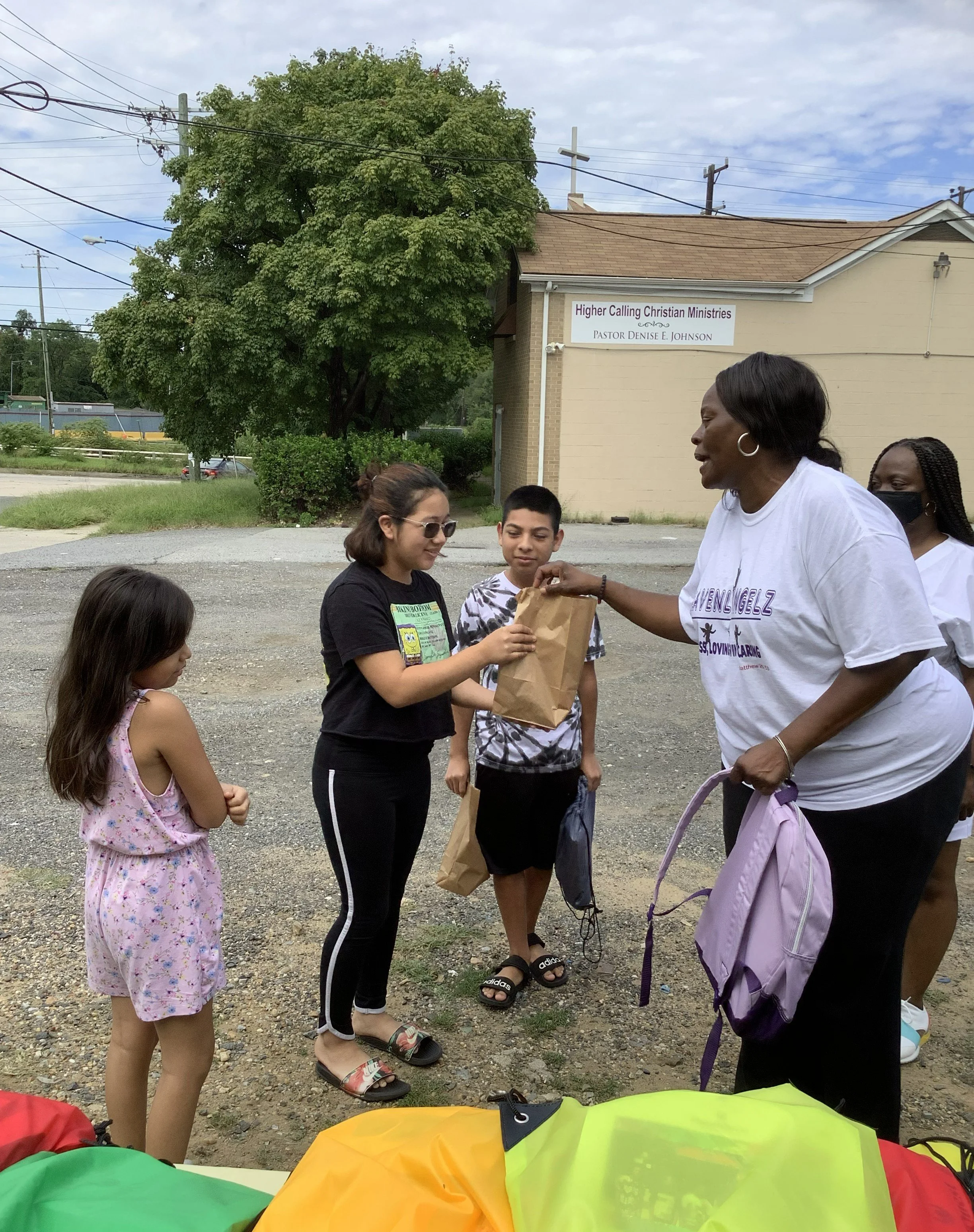 A group of people, including children and adults, standing outdoors near a building with a sign for Higher Calling Christian Ministries, Pastor Denise E. Johnson. The scene appears to be part of a community gathering or distribution event, with colorful items in the foreground and power lines overhead on a partly cloudy day.