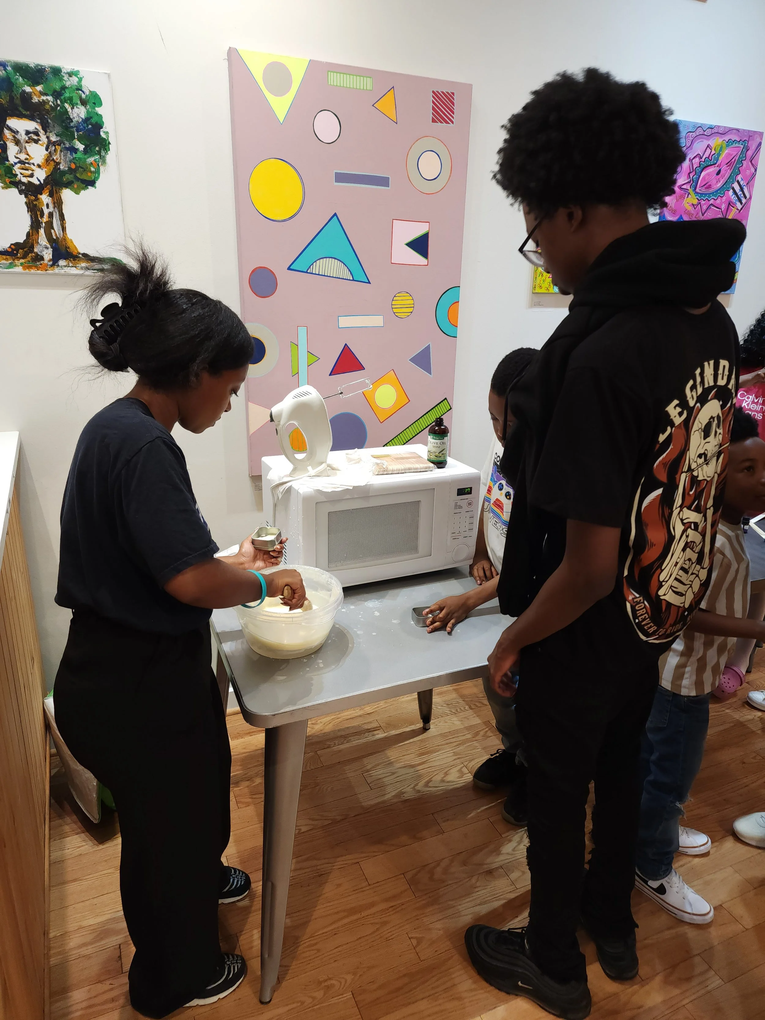 A group of children around a table in an art gallery, with paintings on the wall behind them, and a microwave oven on the table.