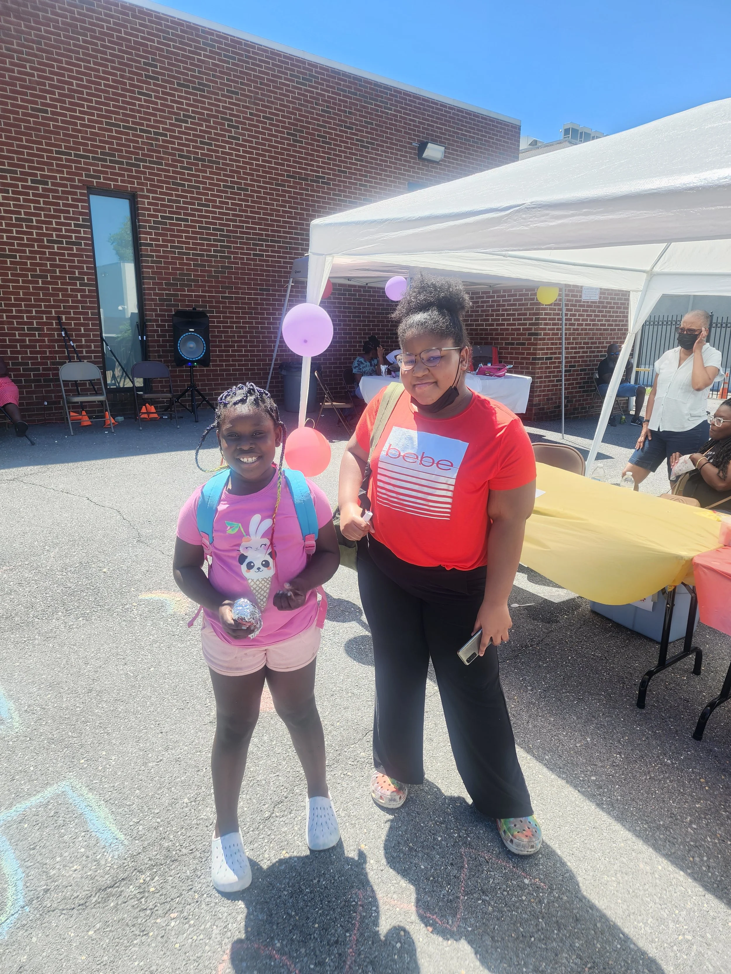 Two girls standing outdoors near a white canopy tent, with balloons and chairs behind them, at a sunny event. One girl is wearing a pink shirt with a bunny graphic and carrying a backpack, and the other is wearing a red shirt with sunglasses and a face mask pulled down.