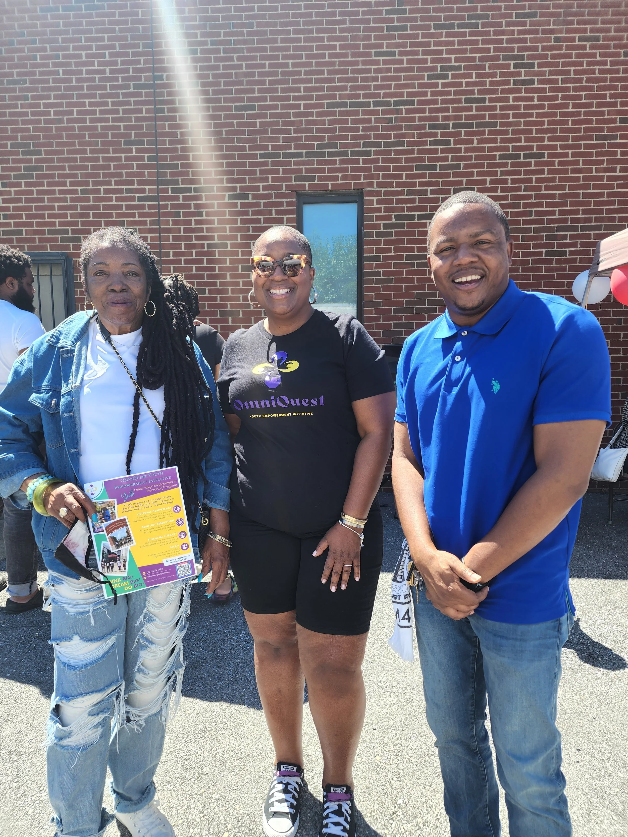 Three people standing outdoors in front of a brick wall, smiling at the camera. The person on the left is a woman with long black dreadlocks, wearing a denim jacket and ripped jeans, holding a colorful flyer. The person in the middle is a woman with 