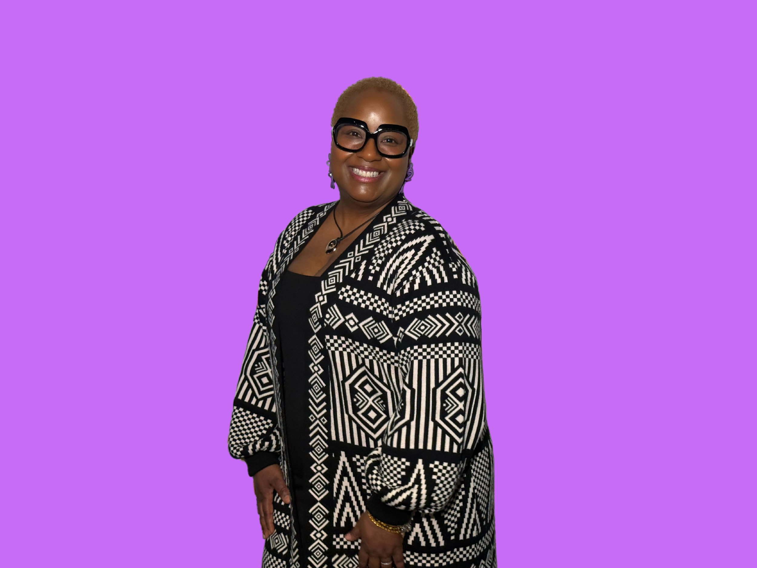A woman with short blonde hair wearing large black glasses, earrings, a black top, and a black-and-white patterned jacket, smiling against a purple background.