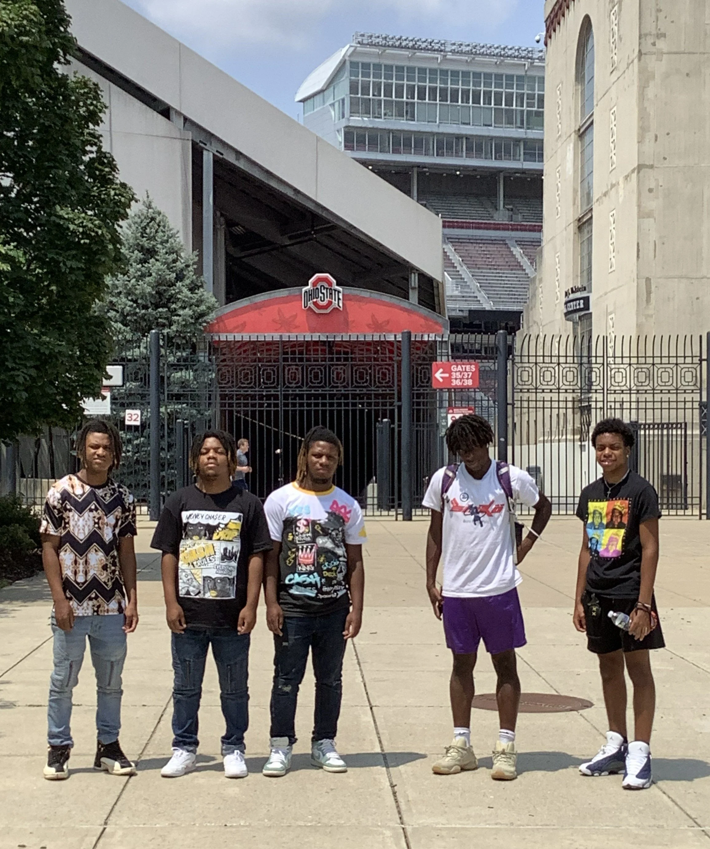 Five young men standing outdoors in front of a closed gate at Ohio State University, with a stadium and school building in the background, on a sunny day.