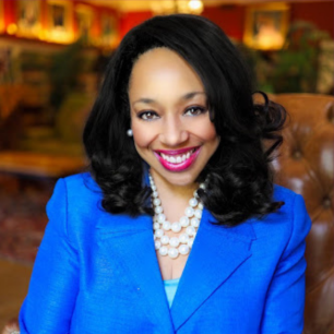 A smiling woman with shoulder-length black hair, wearing a bright blue blazer and a pearl necklace, sitting in a cozy, warmly lit room with wooden furniture and framed pictures on the wall.