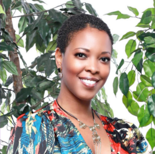 Portrait of a smiling woman with short curly hair, standing in front of leafy green plants, wearing a colorful patterned top and a necklace.