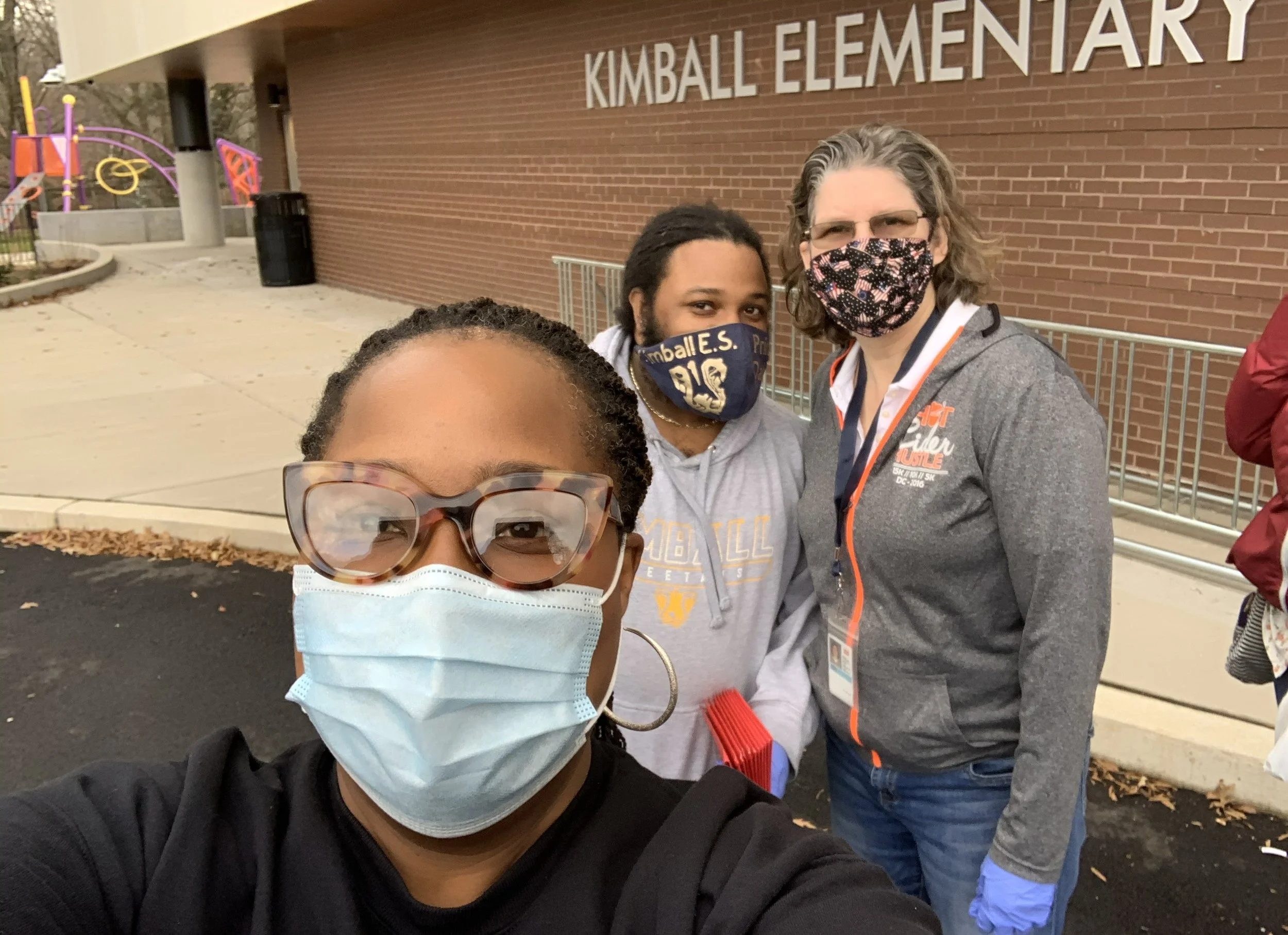 Three people wearing face masks posing for a selfie outside Kimball Elementary School. The person in front is a woman with glasses and a light blue face mask. The middle person is a woman with a colorful face mask and a gray hoodie with orange details. The person on the right is a man with glasses and a black face mask with an American flag design, wearing a gray zip-up jacket.