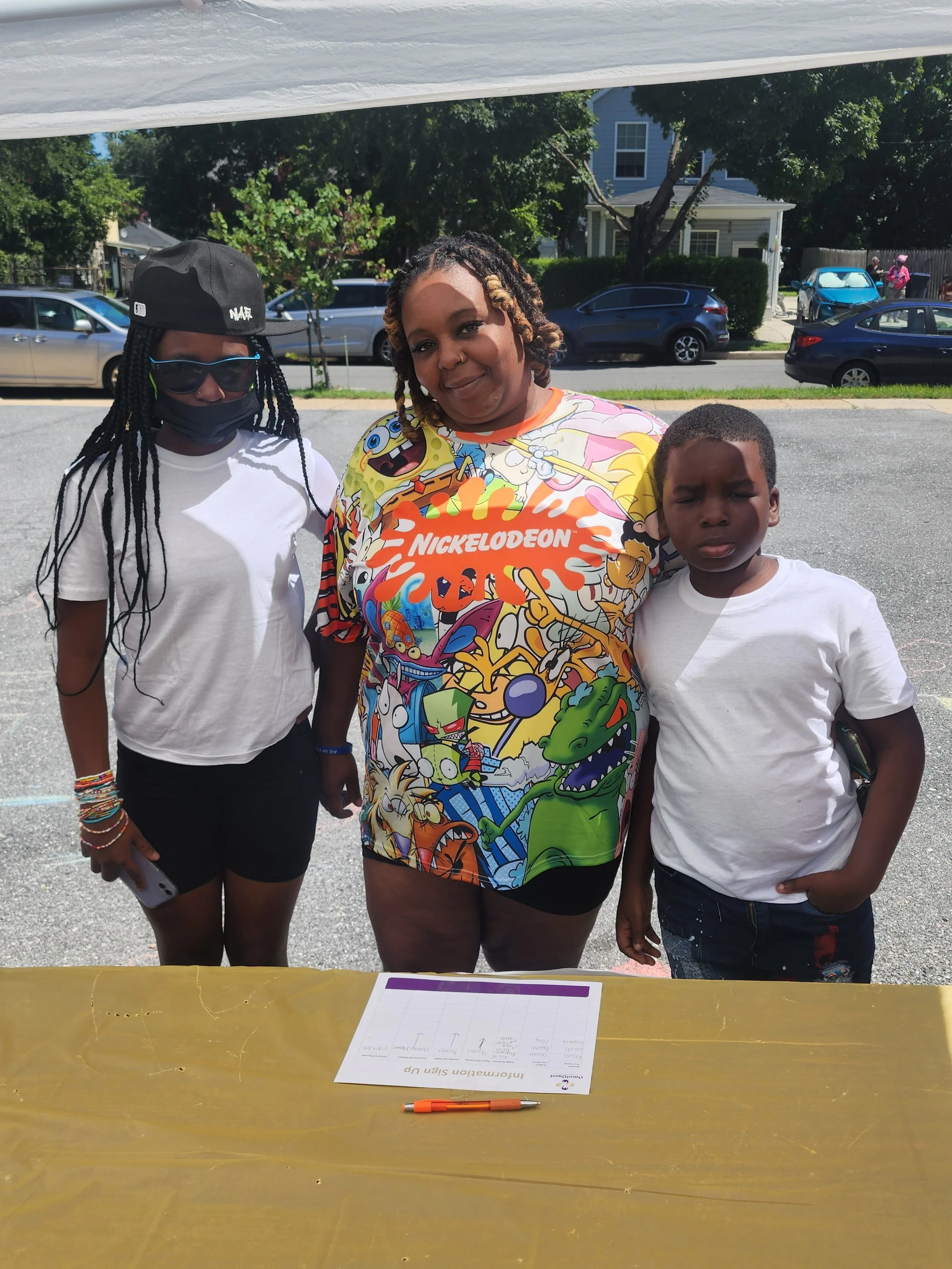 Three people are standing outdoors behind a yellow table with a sign, pen, and papers, with trees, cars, and houses in the background. One woman is wearing a colorful Nickelodeon t-shirt, and two children are standing on each side of her.