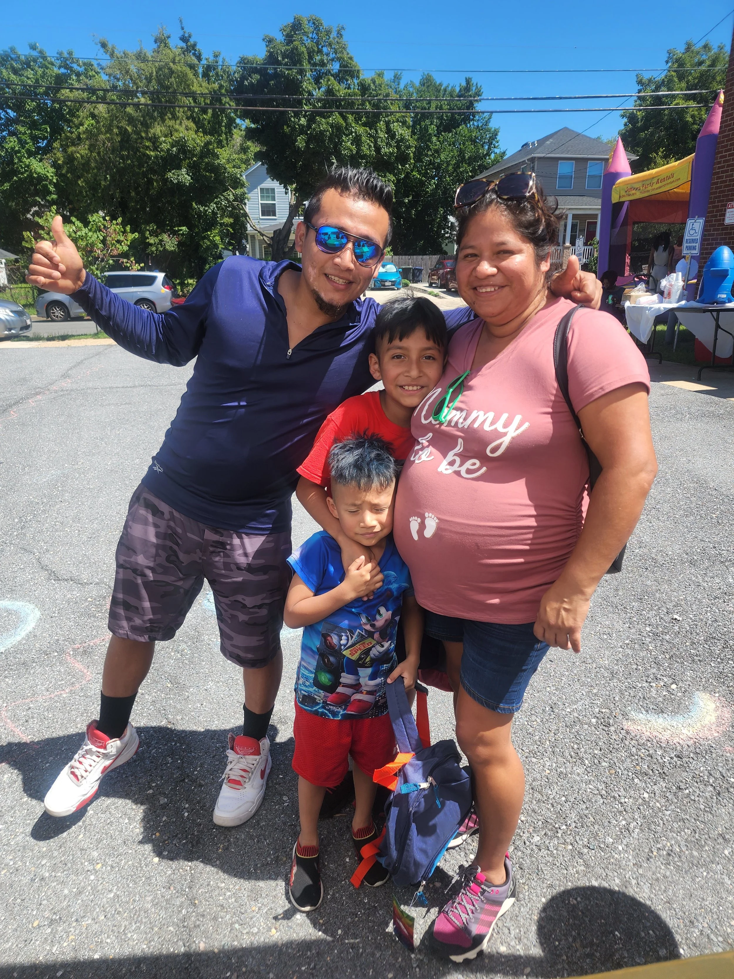 A group of four people, two adults and two children, smiling and posing for a photo outdoors on a sunny day. They are standing on a paved area with trees, houses, and a colorful inflatable bounce house in the background.