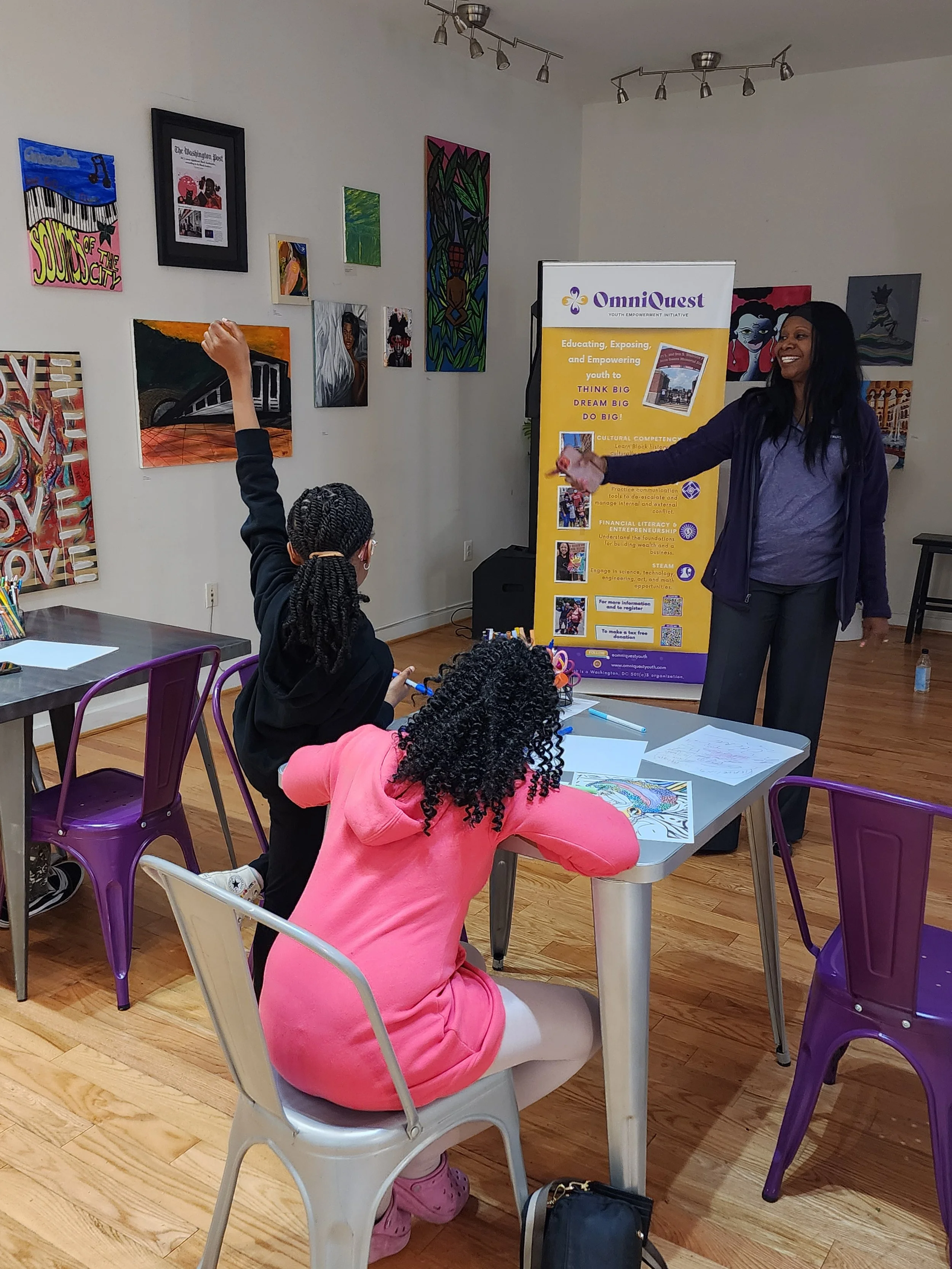 A woman leading a youth workshop in a room decorated with colorful artwork. She is standing near a banner for OmniQuest, talking to young participants seated at tables, some raising their hands.