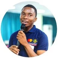 Young man speaking into a microphone, wearing a navy blue polo shirt with logos, in front of a blue curtain.
