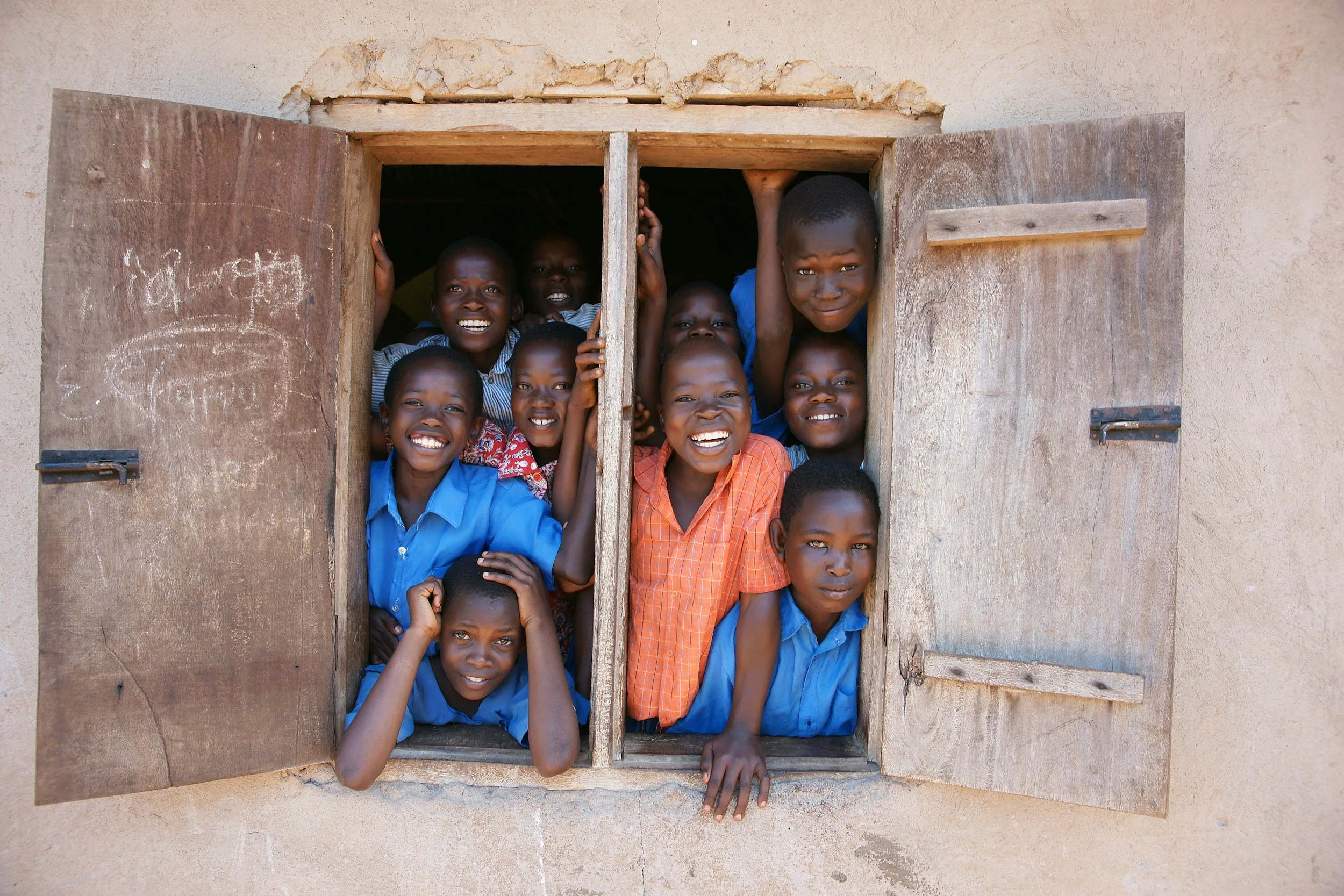 A group of smiling children looking out from a window with open wooden shutters.