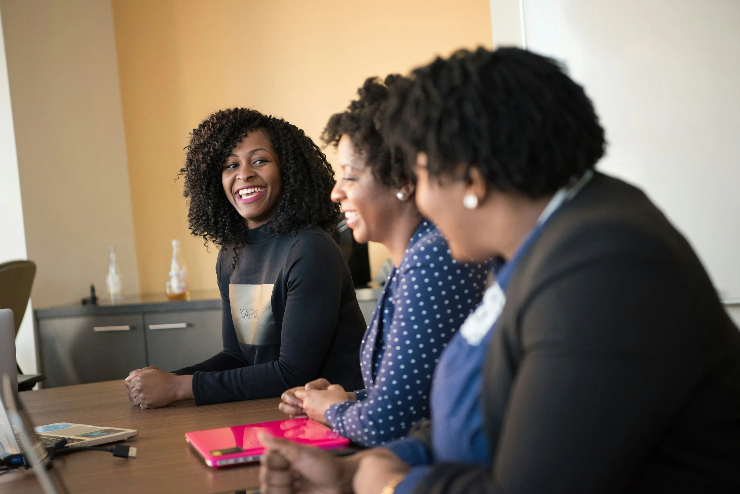 Three women with curly hair seated at a conference table, engaged in a conversation, smiling and appearing happy.