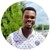 A smiling man standing outdoors with greenery and a cloudy sky in the background.