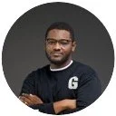 A man with dark skin and short hair, wearing black glasses and a black shirt with a white letter 'G' on the chest, posing with crossed arms against a dark background.