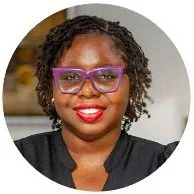 A woman with curly hair, wearing purple glasses and a black top, smiling with red lipstick.