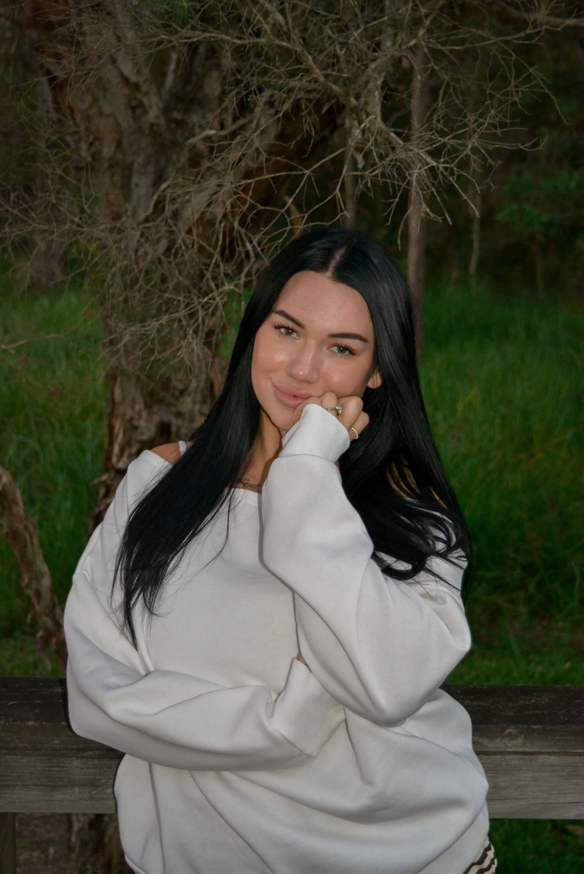 A woman with long black hair and light skin, wearing a white sweatshirt, is sitting outdoors near a wooden railing with a tree with bare branches in the background.
