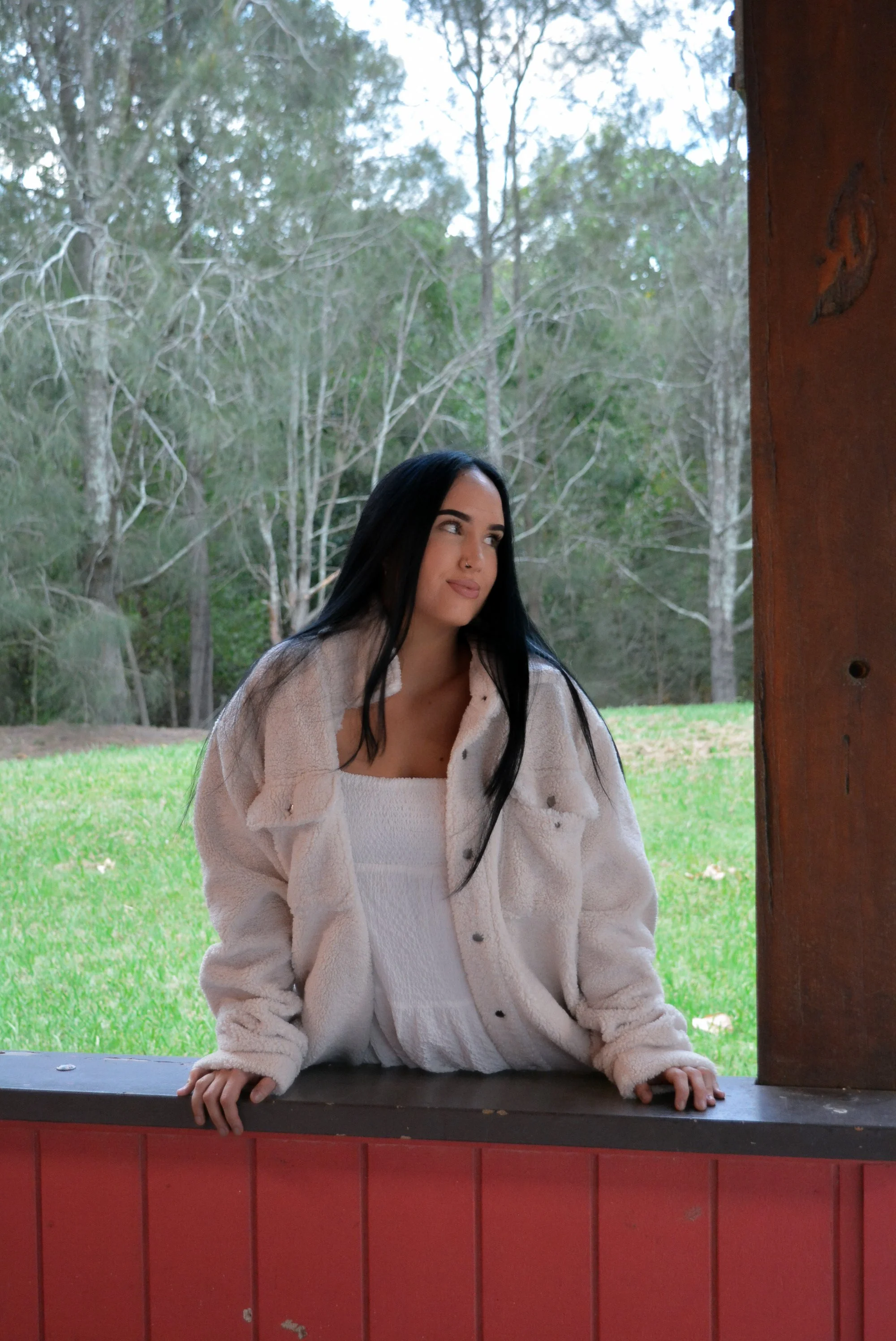 A woman with long black hair wearing a white dress and a cozy beige jacket, leaning on a red wooden ledge outdoors, with a background of trees and a grassy area.