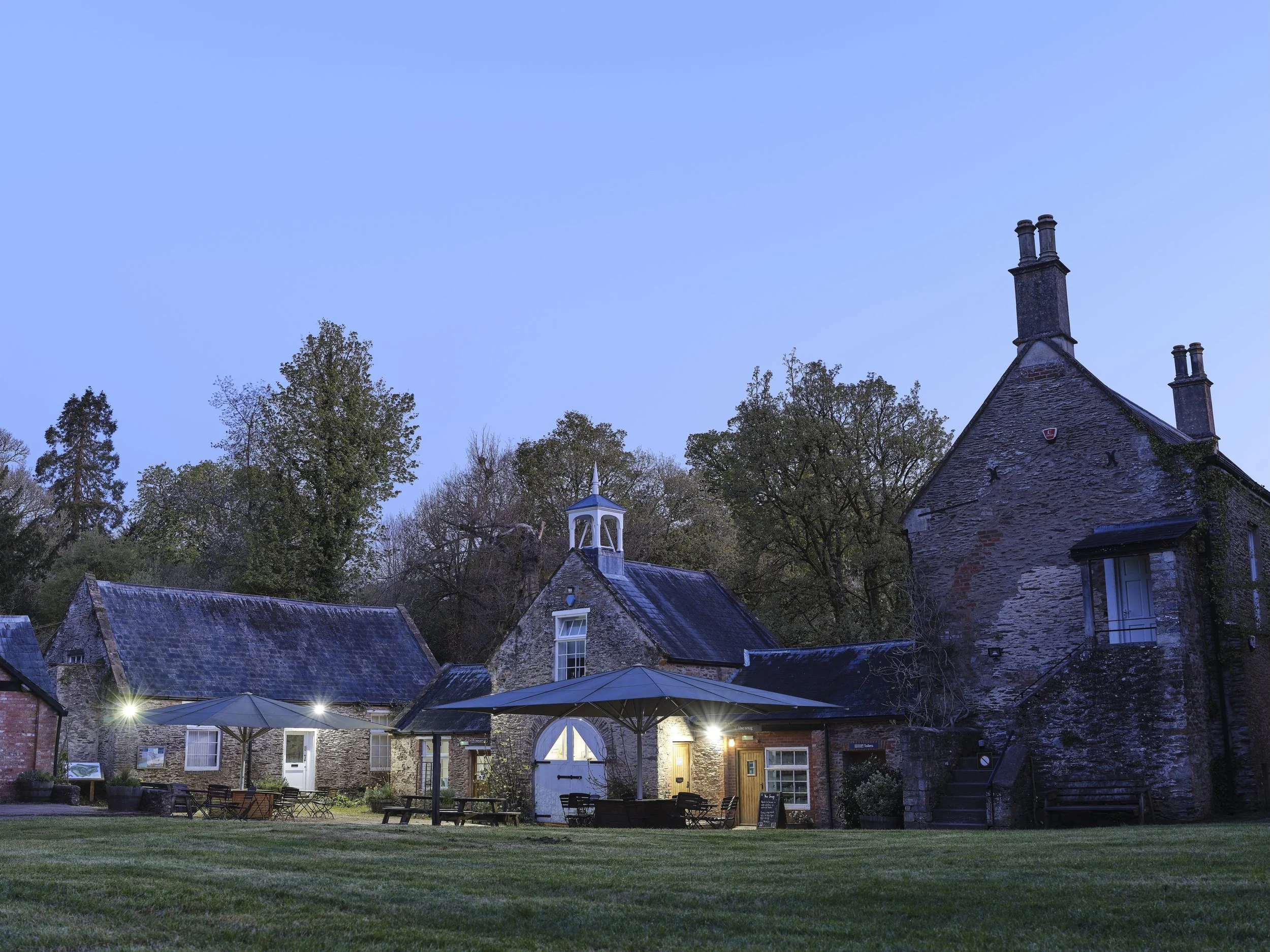 A view of a charming outdoor patio area at dusk, with tables and chairs under umbrellas, situated in front of old stone buildings with pitched roofs and chimneys, surrounded by trees and greenery.