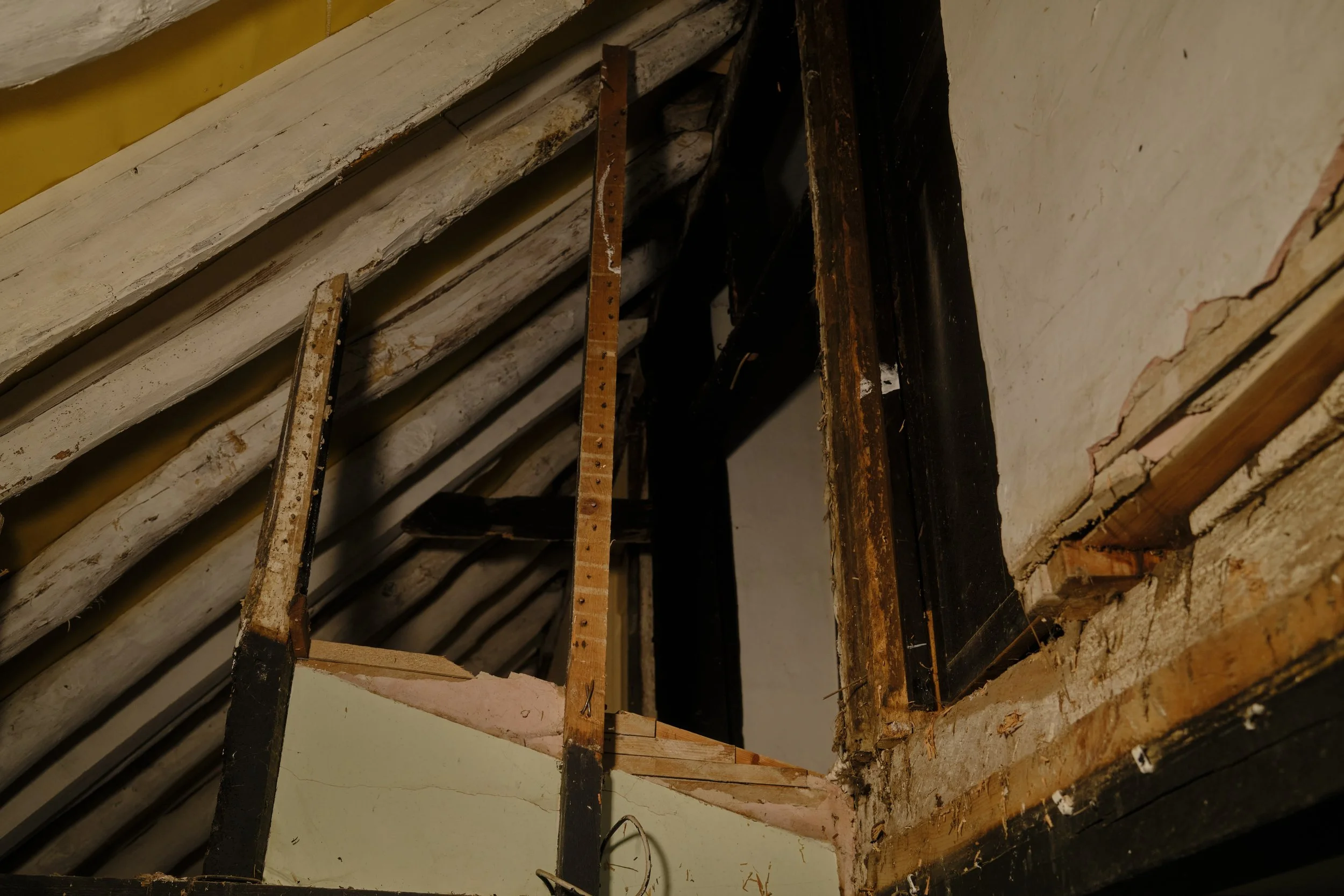 Interior view of a construction site showing wooden and metal framing, with a partially exposed brick wall on the right side, and an unfinished ceiling with wooden beams and insulation.