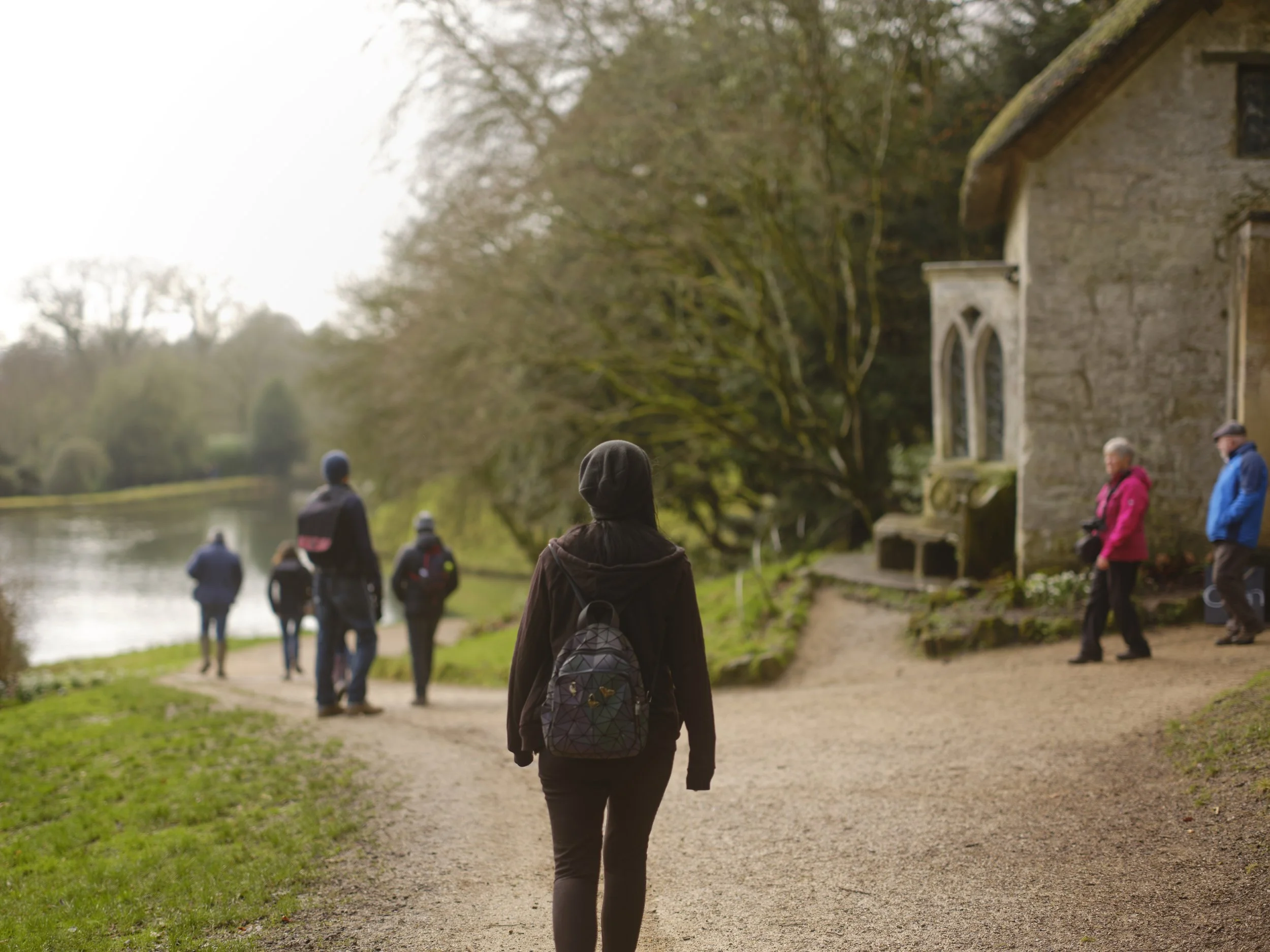 Copyright Laurence Cummins Photography - A woman with a backpack walking on a dirt path along a river, with several other people also walking nearby, and a stone building on the right, trees in the background.