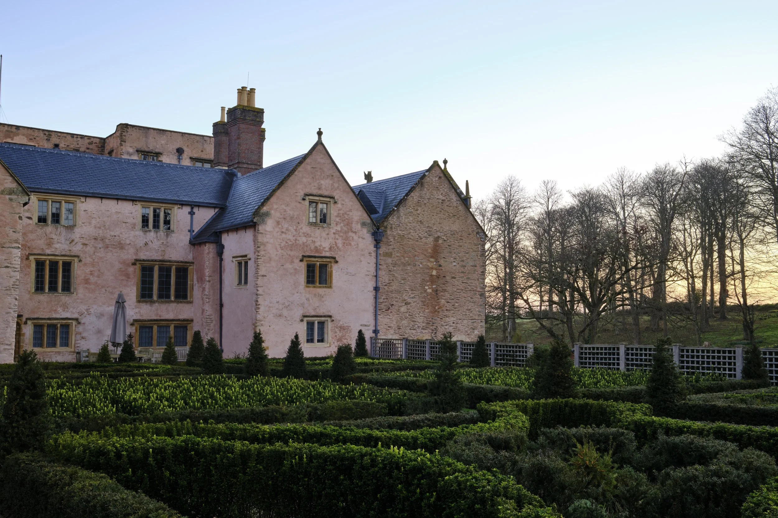 Copyright Laurence Cummins Photography / Halswell Park - A historic pink stone castle with multiple gabled roofs and narrow windows, set against a backdrop of leafless trees at sunset, with a manicured garden in the foreground.