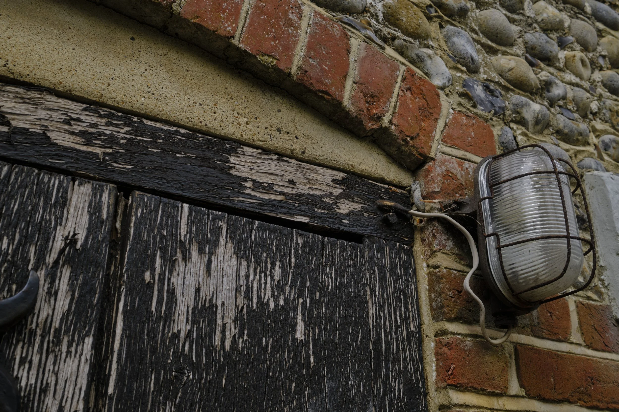 Close-up of a brick and stone exterior wall with a wooden beam and an outdoor light fixture attached by wires.