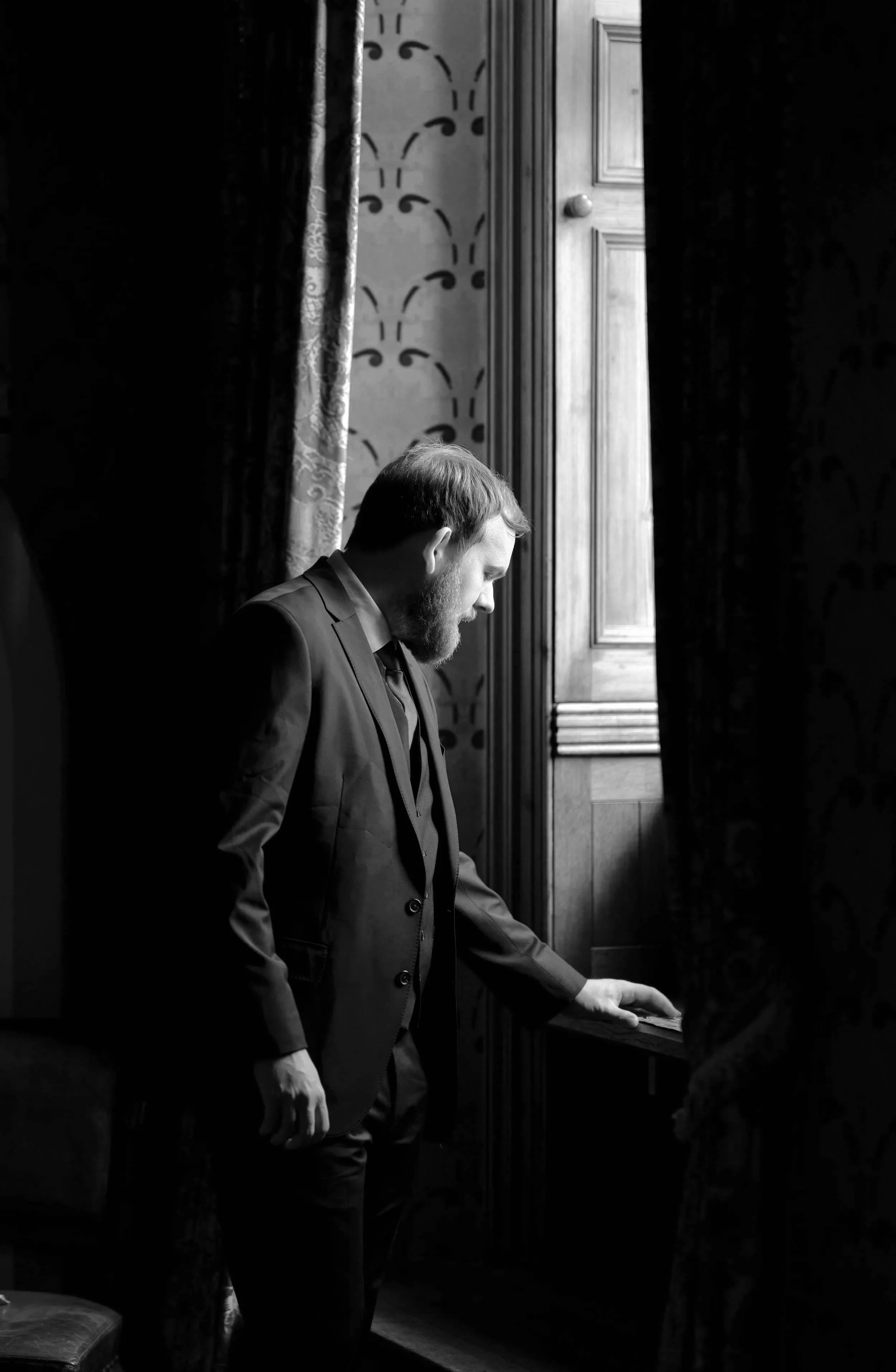 A man in a suit playing a piano in a room with wood-paneled walls and decorative curtains.
