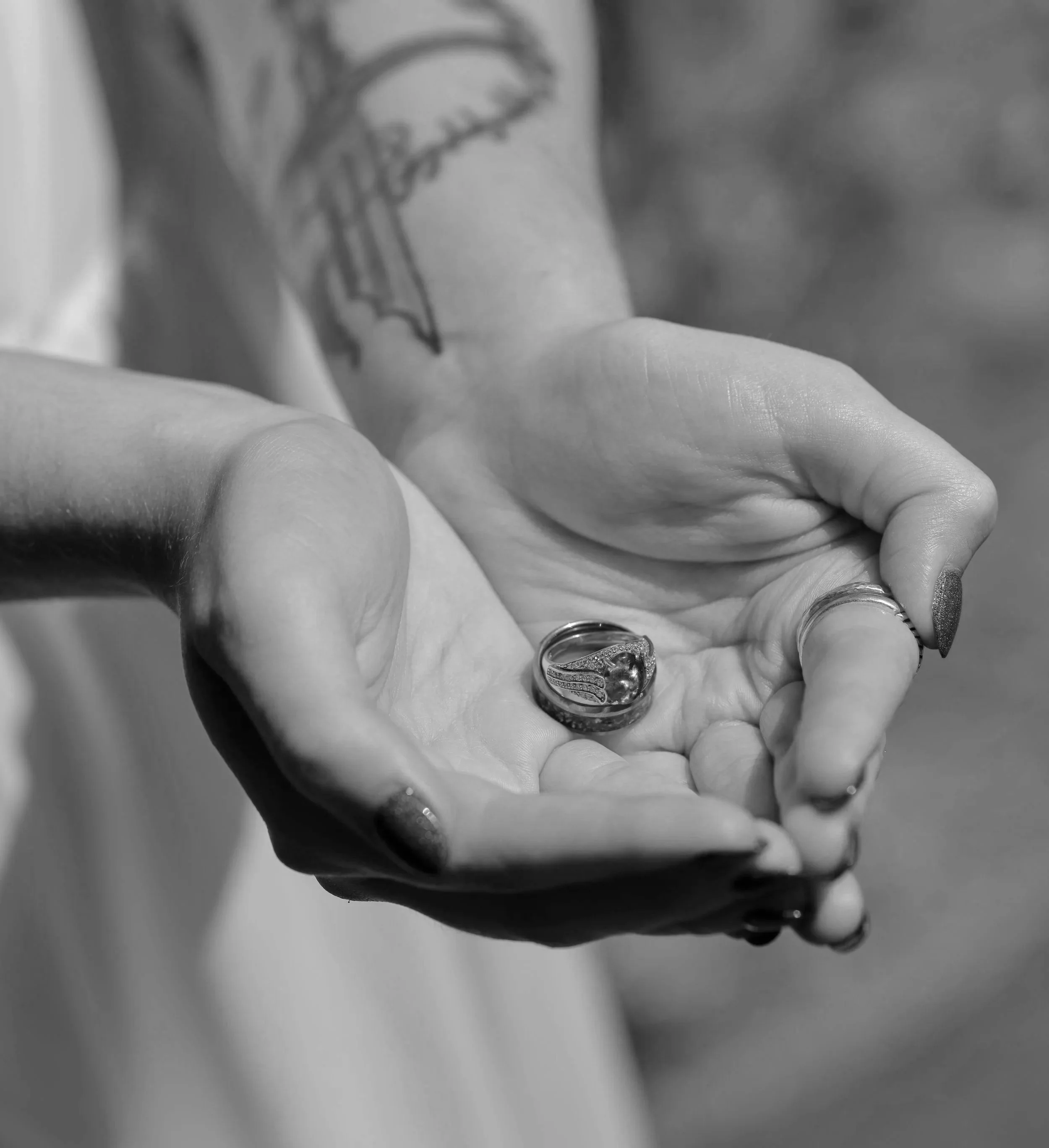 Close-up of a hand wearing multiple rings, including a silver ring with a rainbow-colored opal gemstone, and displaying a tattoo on the wrist, with the background blurred.