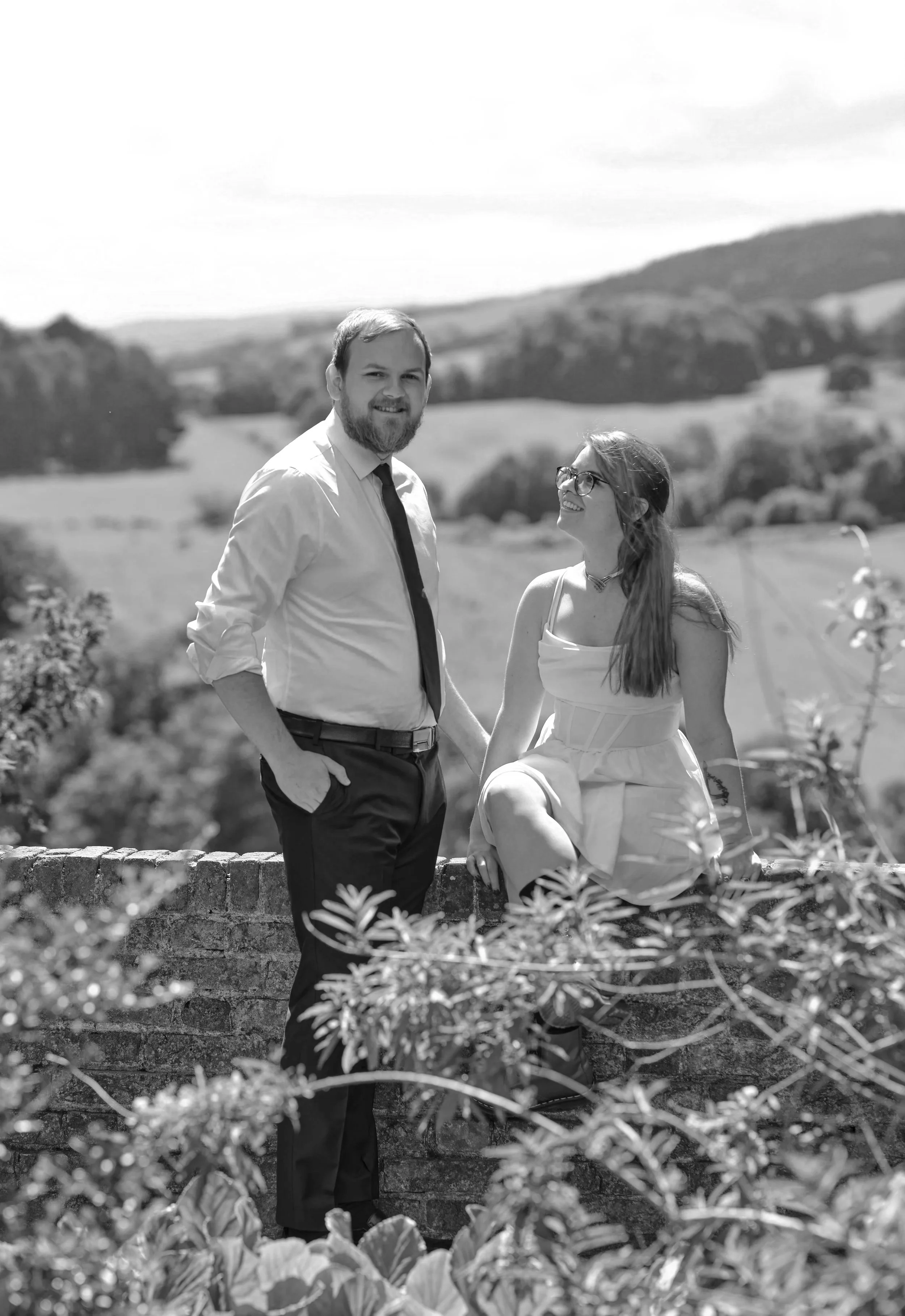 A man in a white shirt and black tie and a woman with blue hair and glasses stand outdoors against a lush green landscape and cloudy sky. The woman is sitting on a brick wall, and they are holding hands.
