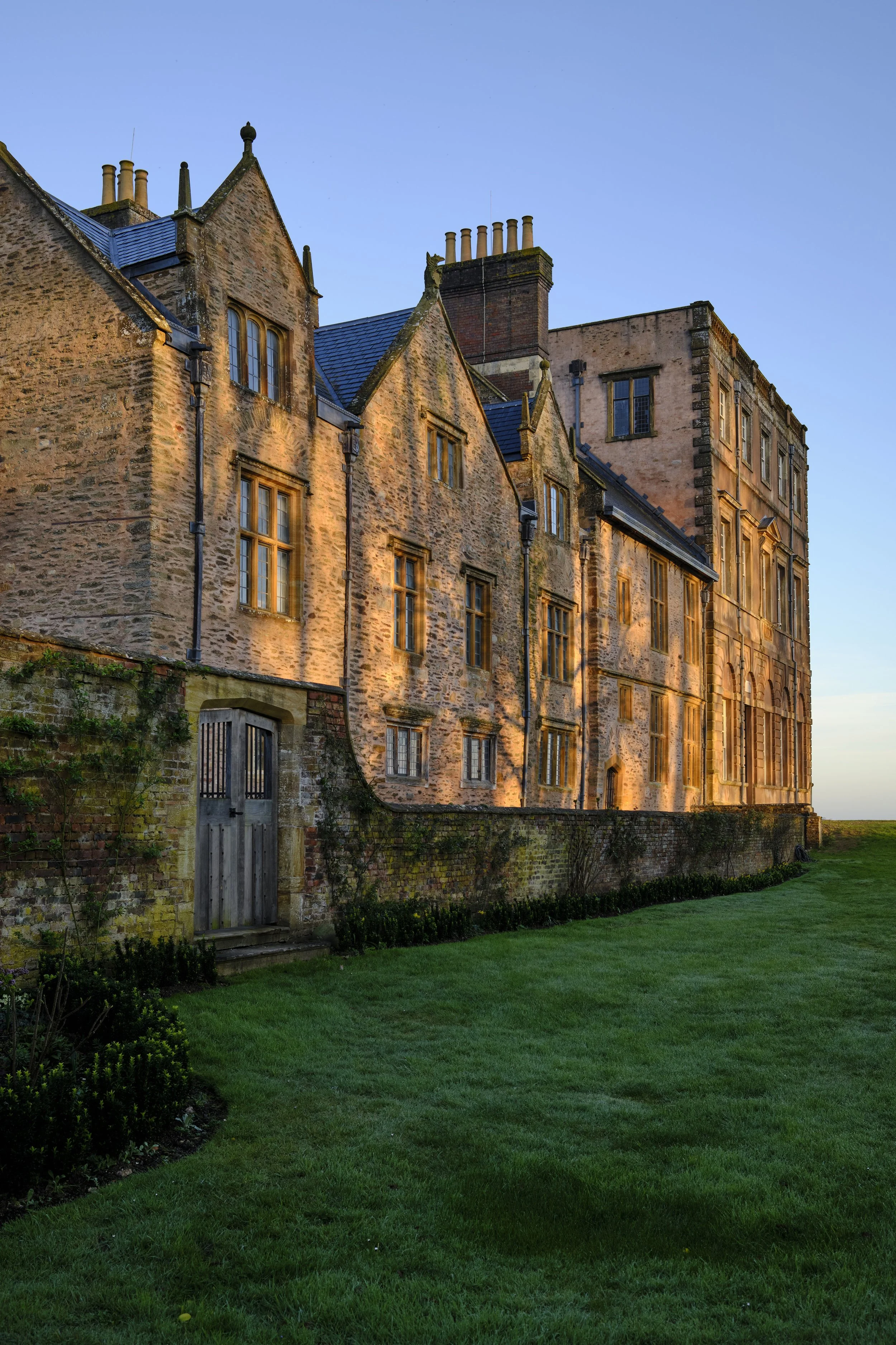 Copyright Laurence Cummins Photography / Halswell Park - An old stone castle with multiple windows and a green lawn in front, bathed in warm sunlight at sunset.