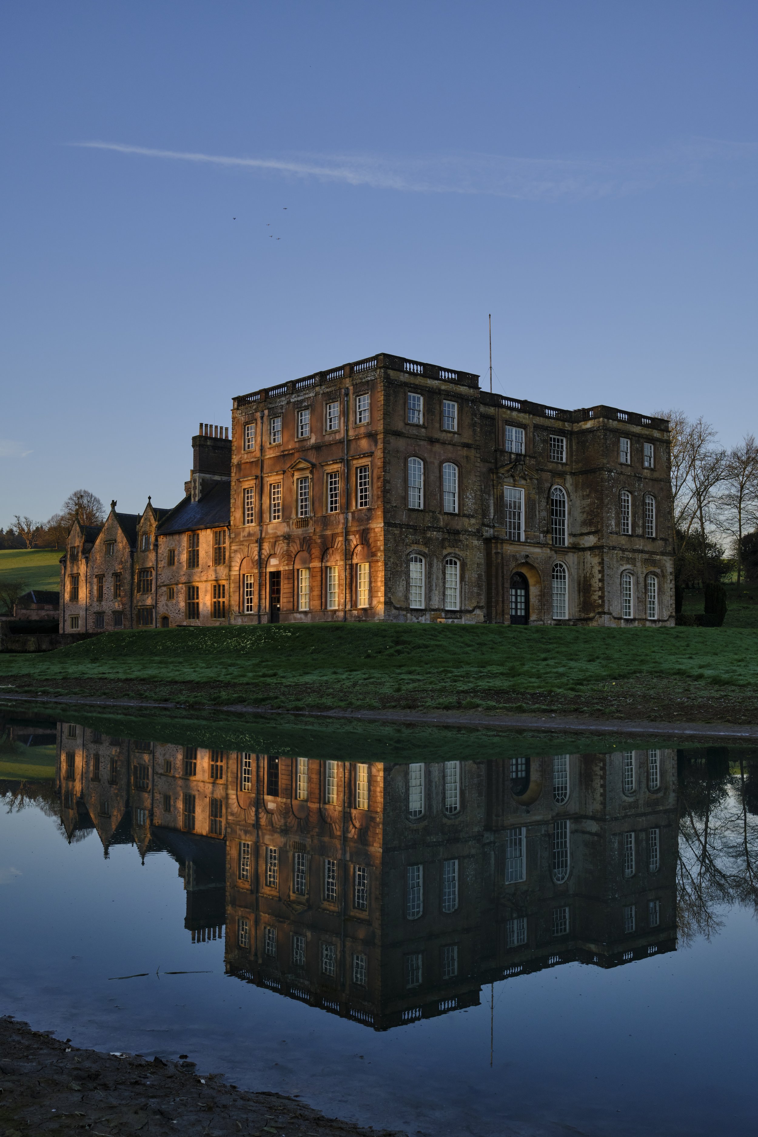 Copyright Laurence Cummins Photography / Halswell Park - A historic castle-like building reflected in a calm body of water under a clear blue sky, with some trees and grassy landscape around.