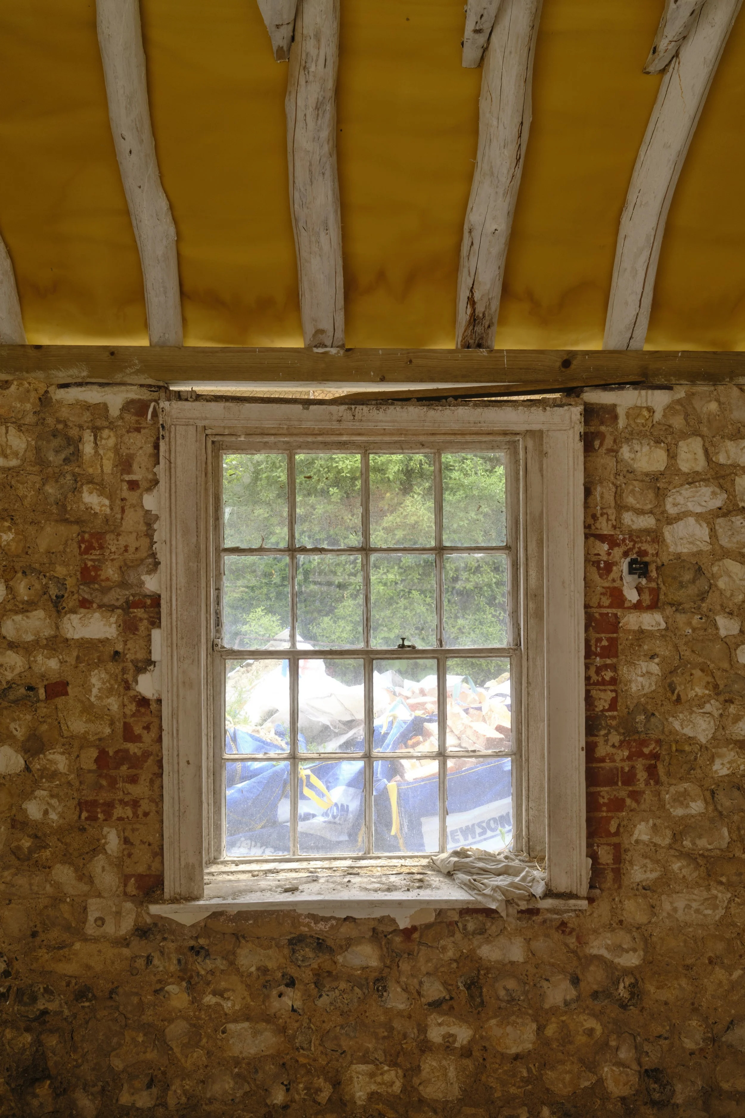 Interior view of a room under renovation with a window showing a view of trees and a construction site outside. The room has a stone wall and an unfinished wooden ceiling with exposed beams.
