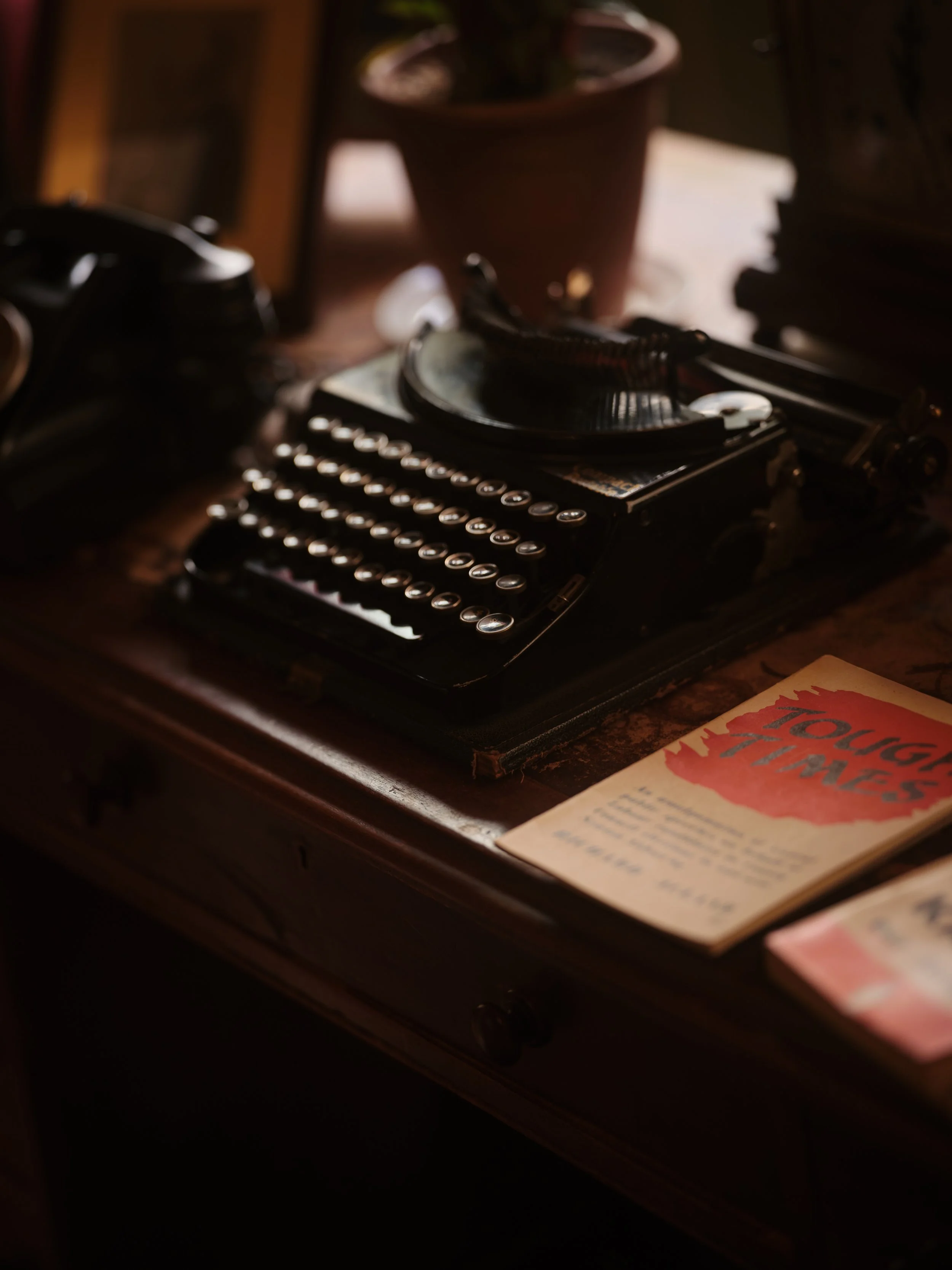 A vintage typewriter with a black and cream color scheme sitting on a wooden desk. Beside it, there is a partially visible book with a red cover and some writing. In the background, a potted plant and some blurred objects are visible.