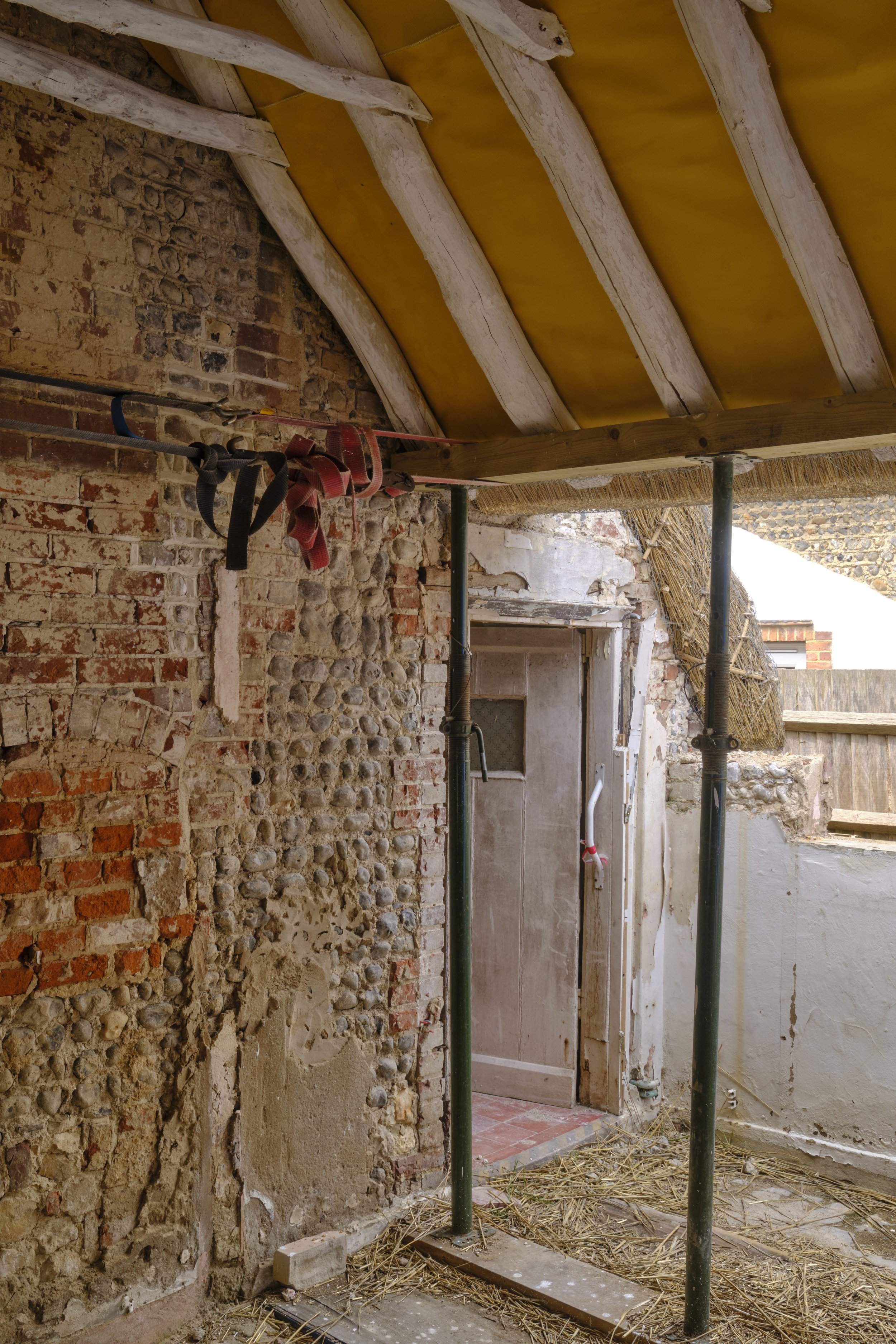 Construction site showing unfinished interior with exposed brick and stone walls, wooden ceiling beams, scaffolding poles, and construction debris, suggesting renovation work.