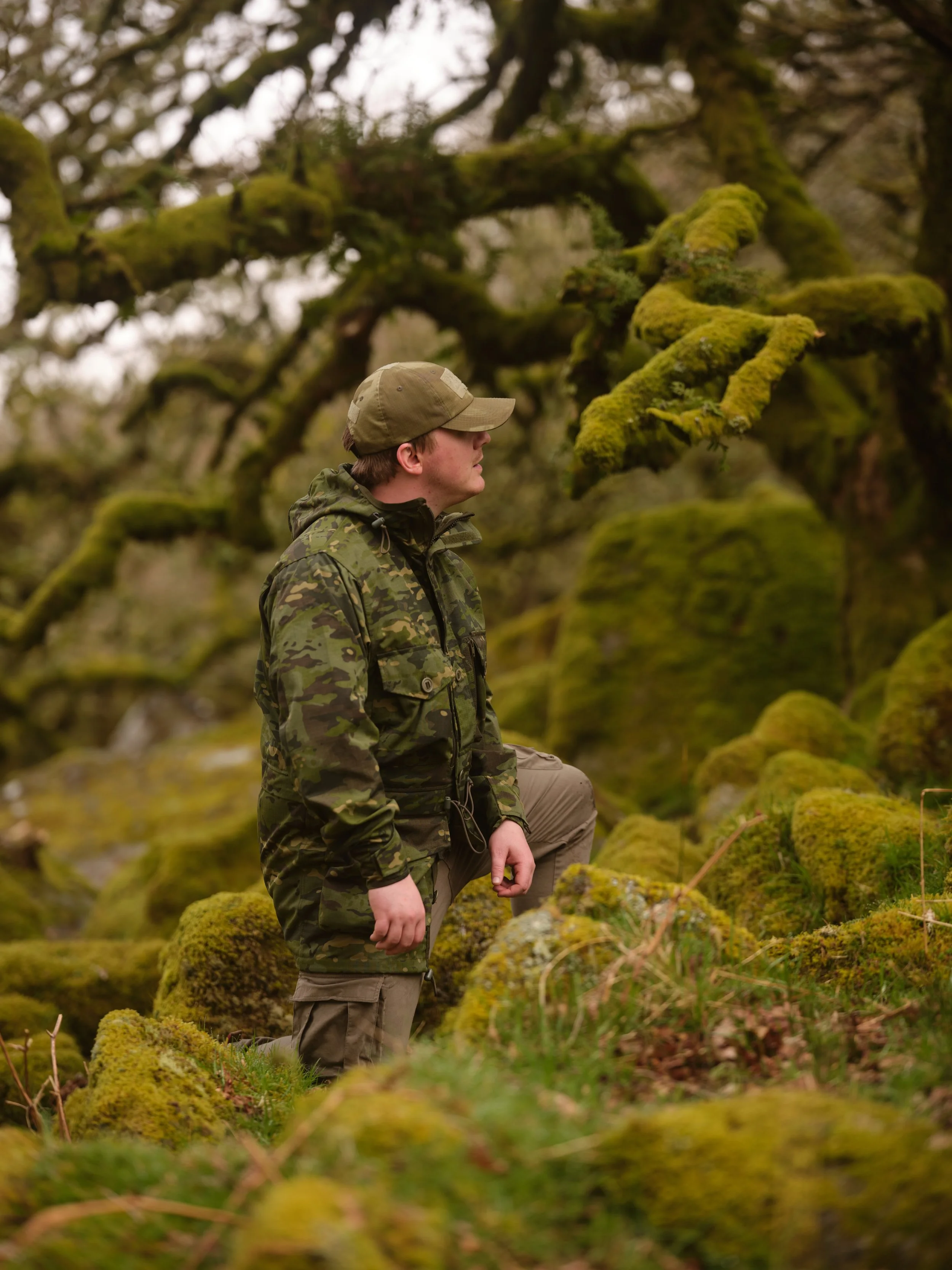 A person dressed in camouflage clothing and a cap, kneeling on one knee in a mossy forest area, surrounded by green moss-covered trees and rocks.