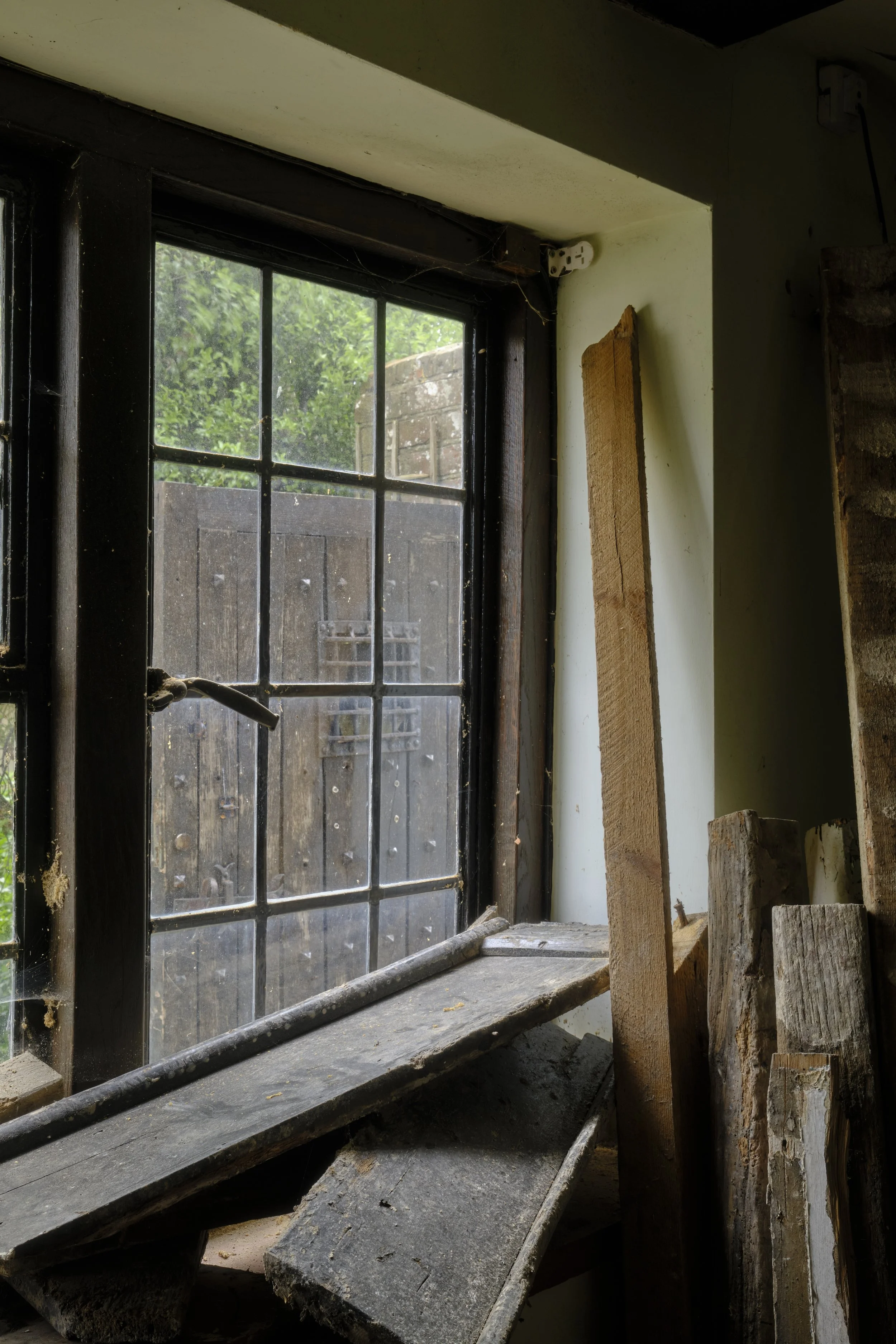 Inside view of a window with dirty, black wooden frames and multiple small panes, with a view of a wooden fence and green foliage outside. The windowsill is cluttered with wood planks, sawdust, and debris.
