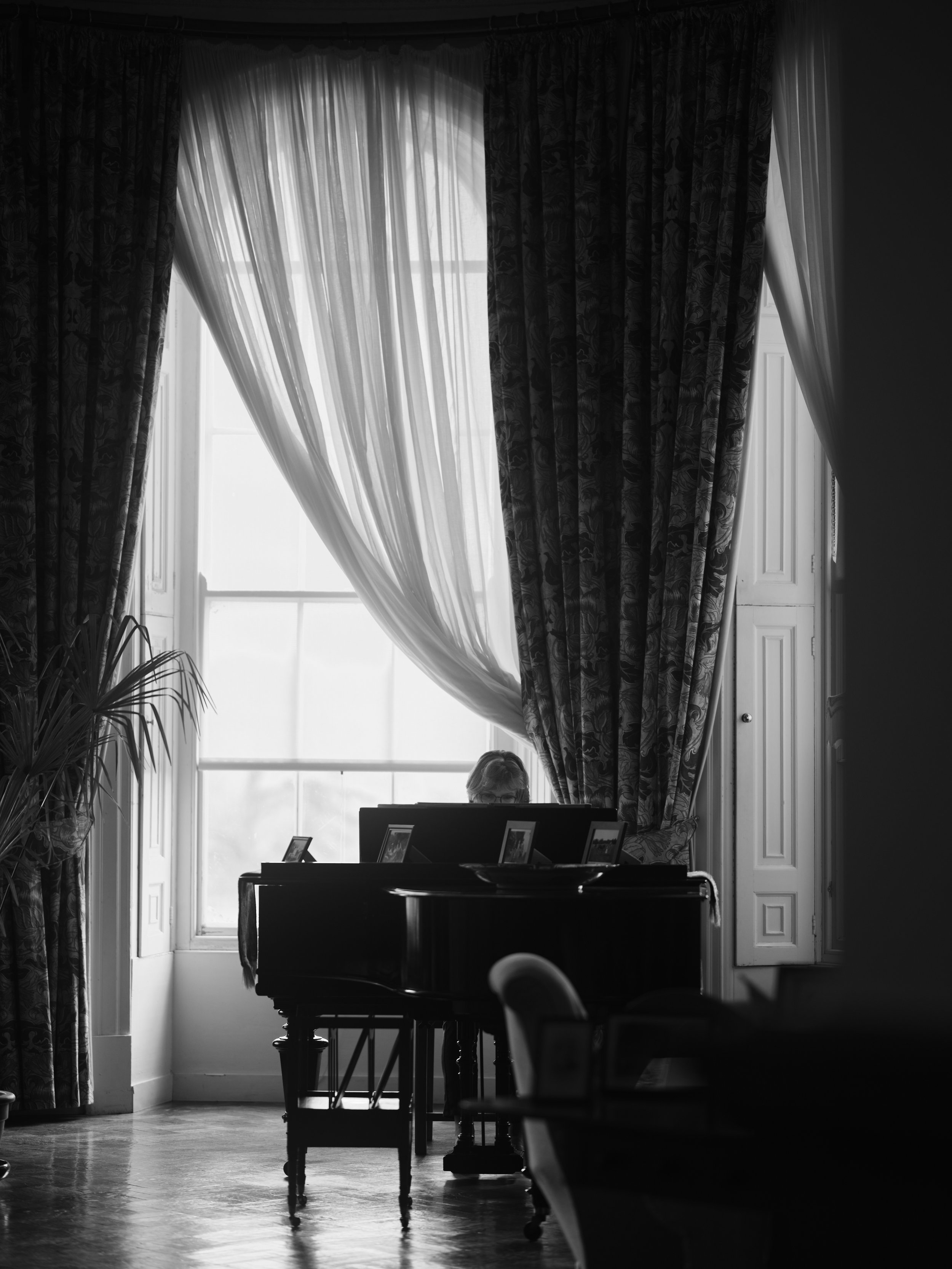 A black and white photo of a woman sitting at a desk by a large window with curtains, with framed photos on the desk and a potted plant nearby.