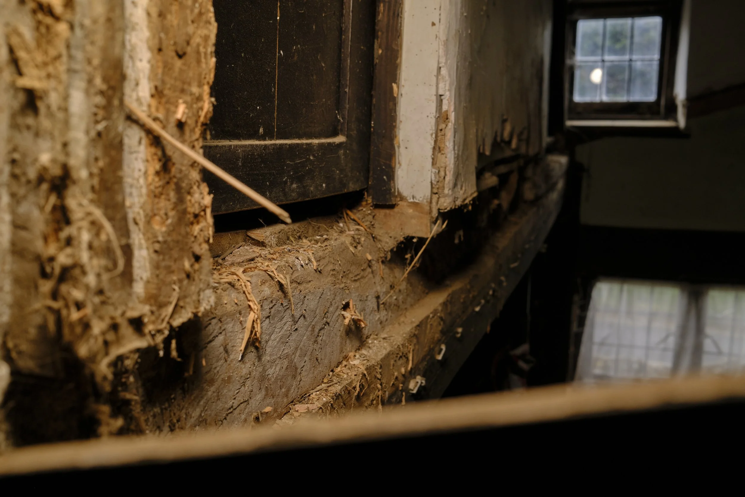 Close-up of a damaged wall with exposed wood and insulation near a window, showing dirt, debris, and structural deterioration.