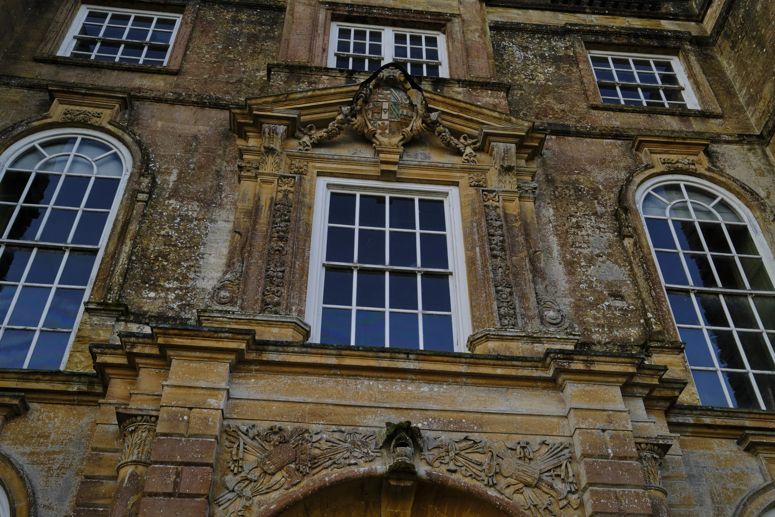 Copyright Laurence Cummins Photography / Halswell Park - Close-up of an old, ornate building facade with three large windows and intricate stone carvings around the middle window, including a heraldic shield at the top.