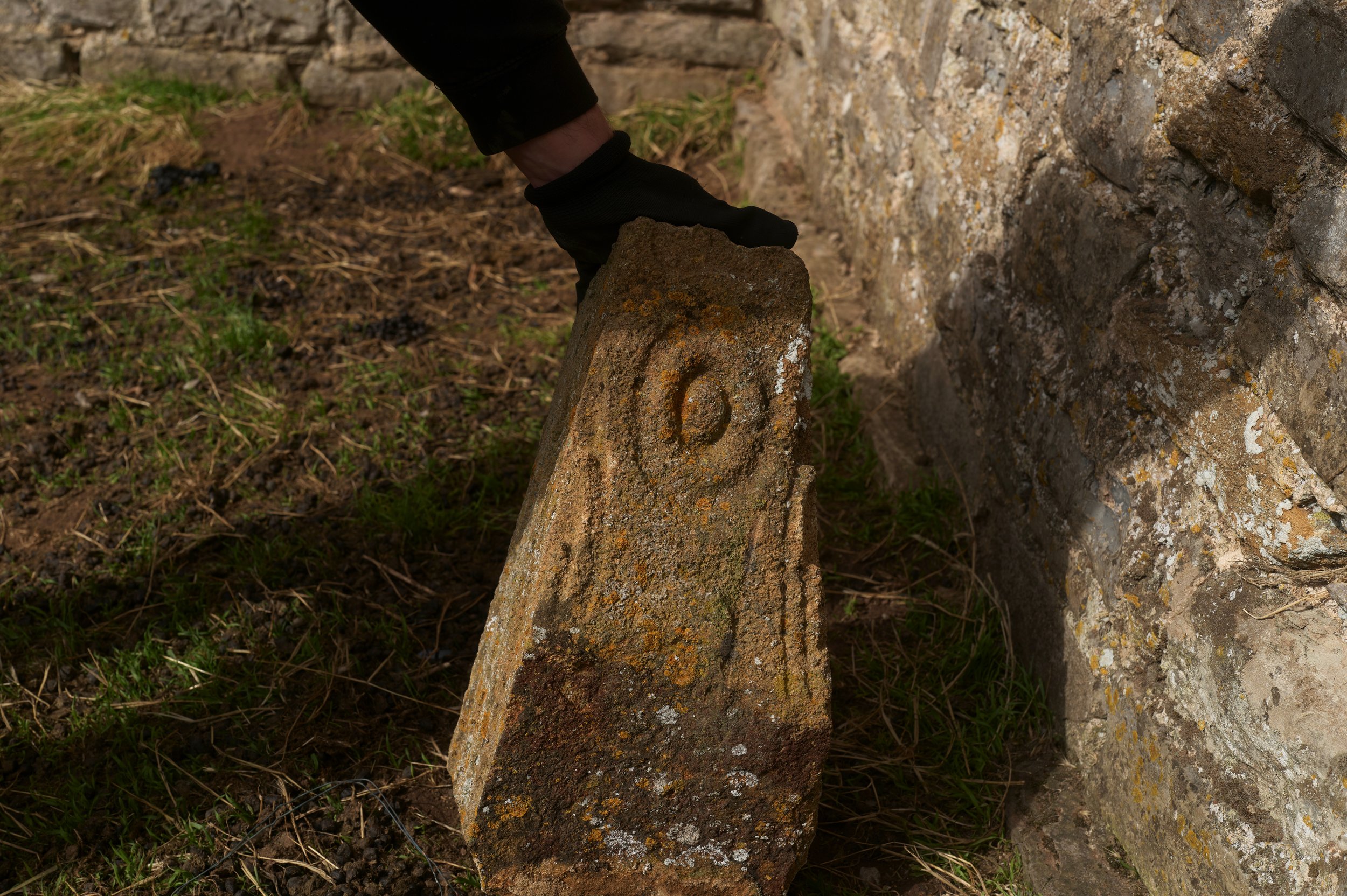 Person wearing black gloves placing a weathered brick on the ground near an old stone wall.