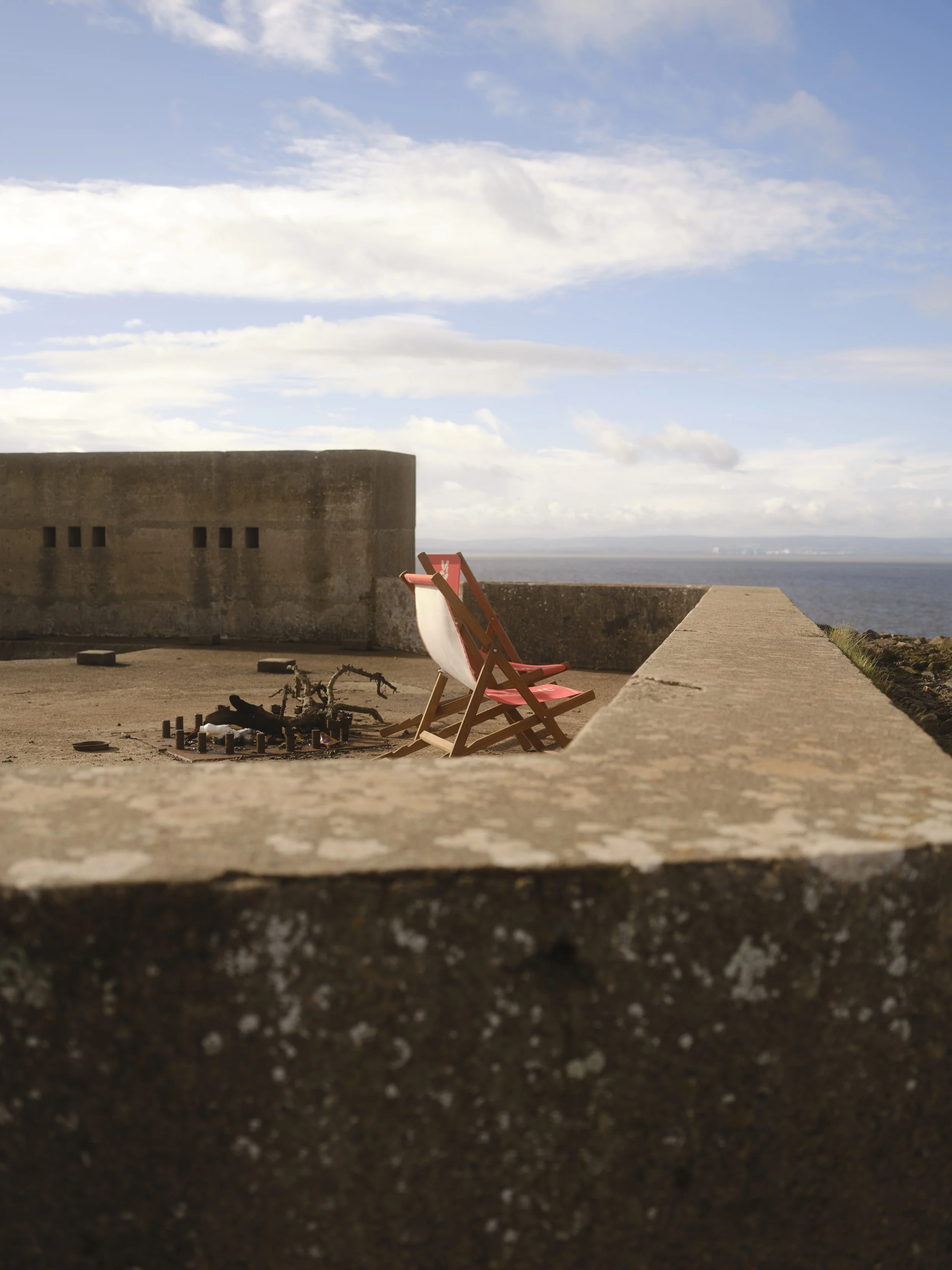 A concrete ledge overlooking the ocean under a partly cloudy sky, with a red and white deck chair and a small fire pit with ashes nearby.