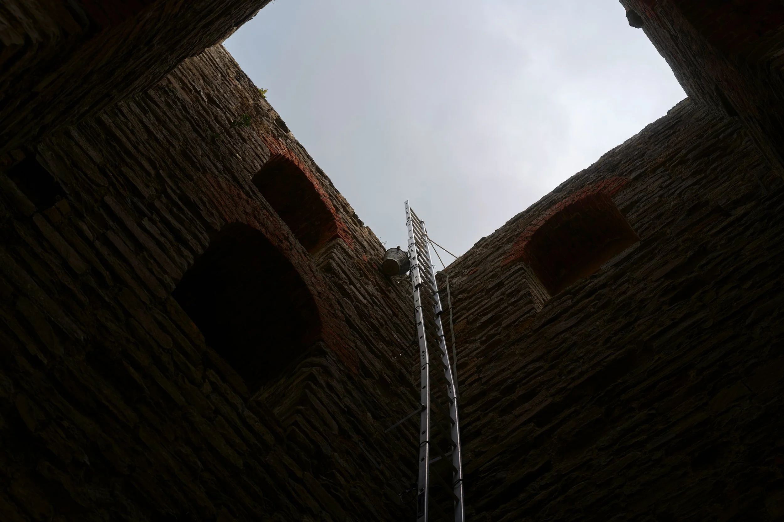 Looking up from the bottom of a brick well or chimney, showing the brick walls and a metal ladder leading up toward the opening at the top, with the sky visible.